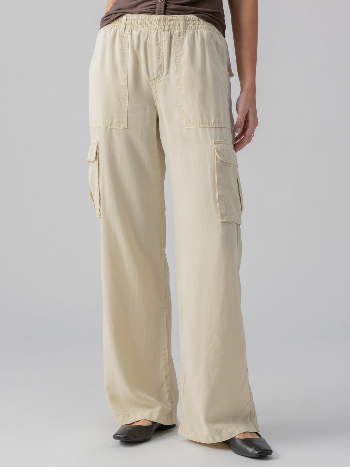 A person is dressed in Sanctuary Clothing's Relaxed Reissue Birch cargo pants, featuring large side pockets, paired with a dark brown top. They are wearing black, pointed-toe flats. The photo captures the outfit from the waist down in front of a plain, light gray background.