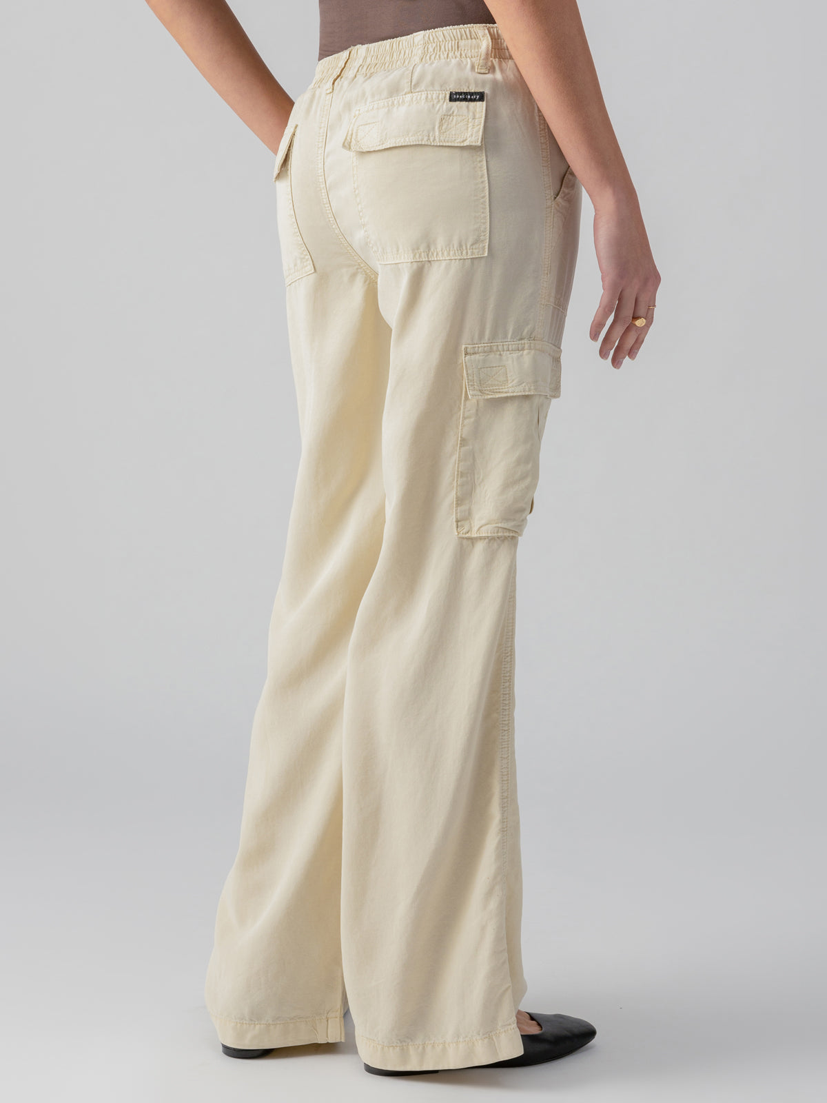 A person wearing the Relaxed Reissue Birch from Sanctuary Clothing, which are high-waisted, wide-leg beige cargo pants with multiple pockets, stands with their back to the camera. They are also sporting a brown top and black shoes, with a ring on their left hand. The background is a plain light gray.