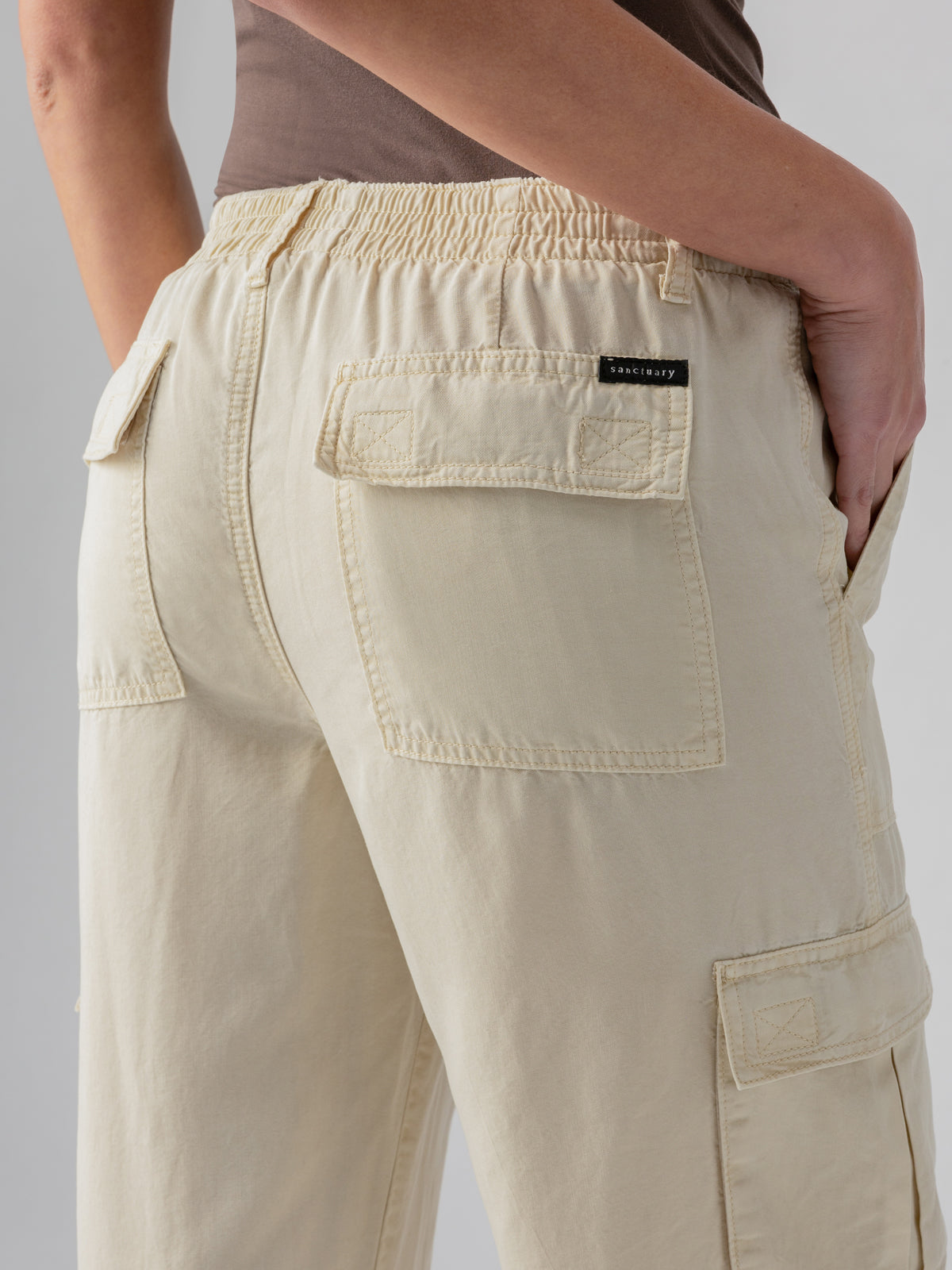 A close-up of a person wearing the Relaxed Reissue Birch beige cargo pants by Sanctuary Clothing, featuring an elastic waistband and multiple pockets, including two large square pockets on the back. The person’s hand rests on their hip, and they are wearing a brown top. The brand tag reads "Sanctuary.