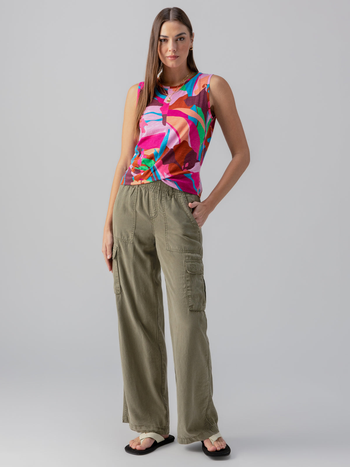 A woman stands confidently with one hand on her hip, wearing a vibrant abstract-patterned sleeveless top paired with Sanctuary Clothing's Relaxed Reissue Burnt Olive cargo pants. She has straight, long brown hair and is sporting black flip-flops against a plain gray background.