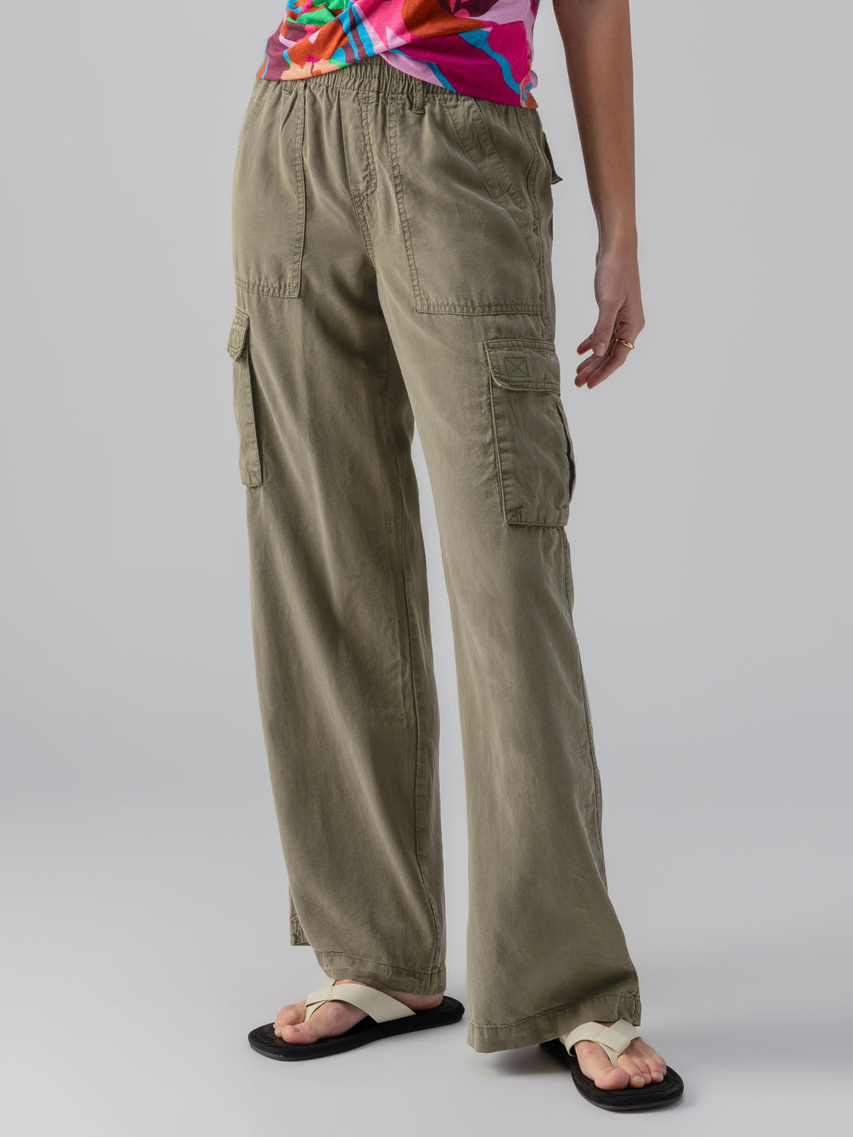 A person wearing the Relaxed Reissue Burnt Olive cargo pants by Sanctuary Clothing and a colorful shirt stands with one hand by their side. Their feet are clad in white sandals with black soles. The background is plain gray.