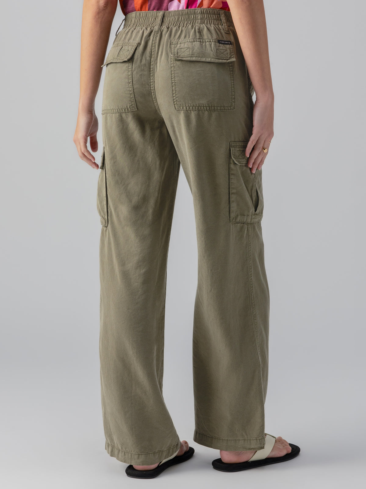 A person is shown from the back, wearing the Sanctuary Clothing Relaxed Reissue Burnt Olive pants, which are loose-fitting cargo pants with multiple pockets. Paired with black sandals, these pants feature an elastic waistband and flap pockets on the back and sides. The background is plain and light gray.