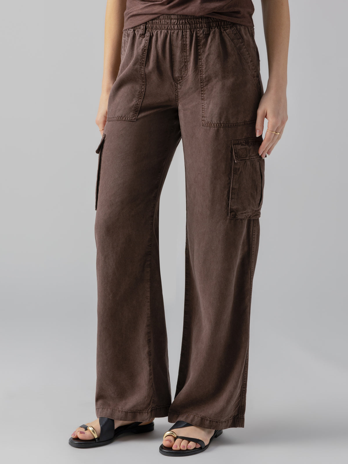 A person is modeling the Sanctuary Clothing's Relaxed Reissue Mud Bath ensemble, featuring a dark brown sleeveless top paired seamlessly with matching wide-leg cargo pants that have multiple pockets. Their right hand rests casually in one of the pockets while their left hand hangs naturally by their side. Complementing the outfit are black sandals adorned with a gold buckle. They stand against a plain light gray background.