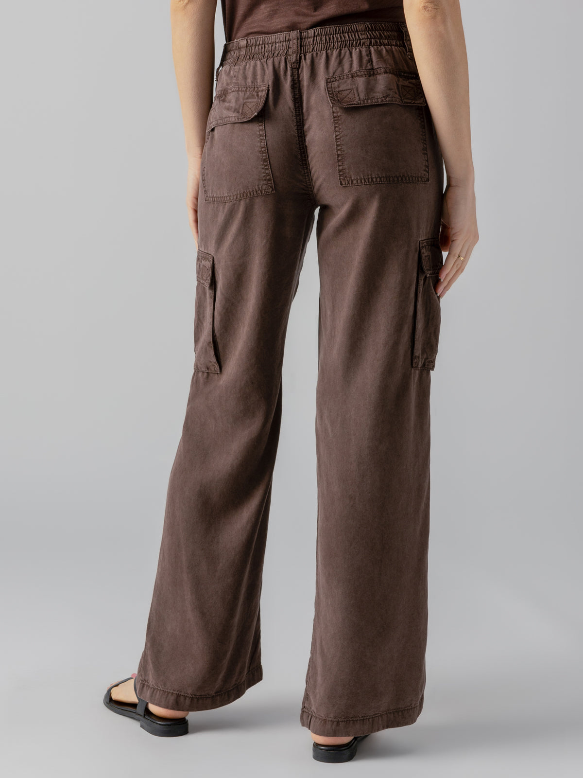 A person stands facing away, showcasing the Relaxed Reissue Mud Bath wide-leg cargo pants from Sanctuary Clothing, featuring flap pockets on the back and sides. The pants are designed with an elastic waistband. The person is wearing open-toed black sandals against a plain light gray background.