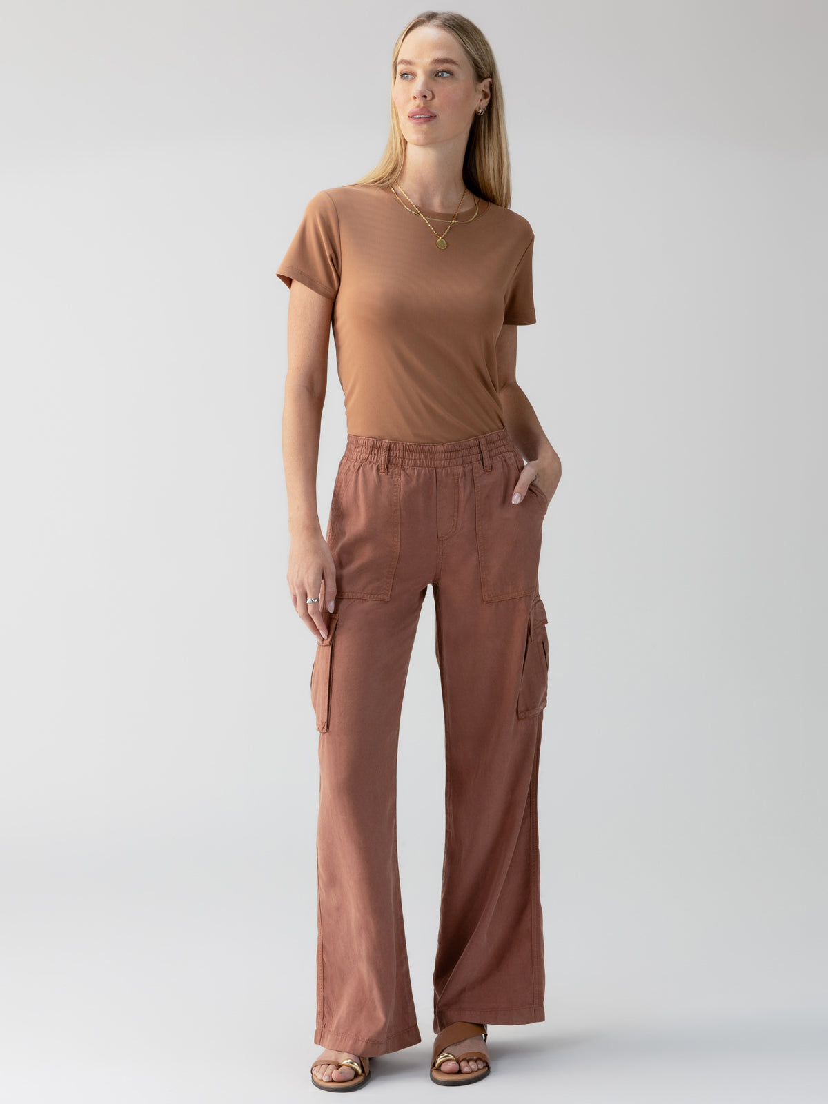 A woman stands against a plain backdrop wearing the Relaxed Reissue Washed Clay short-sleeved top by Sanctuary Clothing, paired with brown cargo pants featuring pockets and sandals. She has one hand in a pocket and gazes slightly to the side, posing in a laid-back manner.