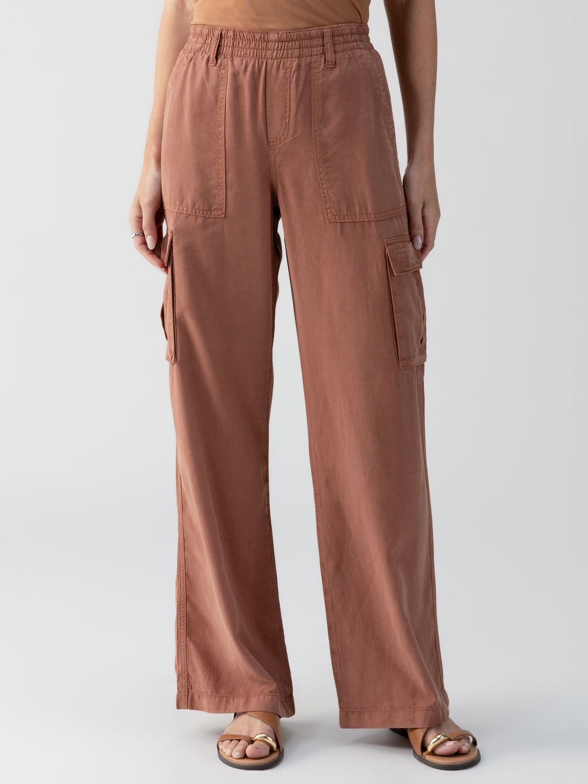 A person wearing Sanctuary Clothing's Relaxed Reissue Washed Clay high-waisted, loose-fitting cargo pants in a rusty brown color. The pants feature multiple pockets, including large flap-covered pockets on the sides. The person is also sporting a tucked-in top and open-toed sandals with gold accents.