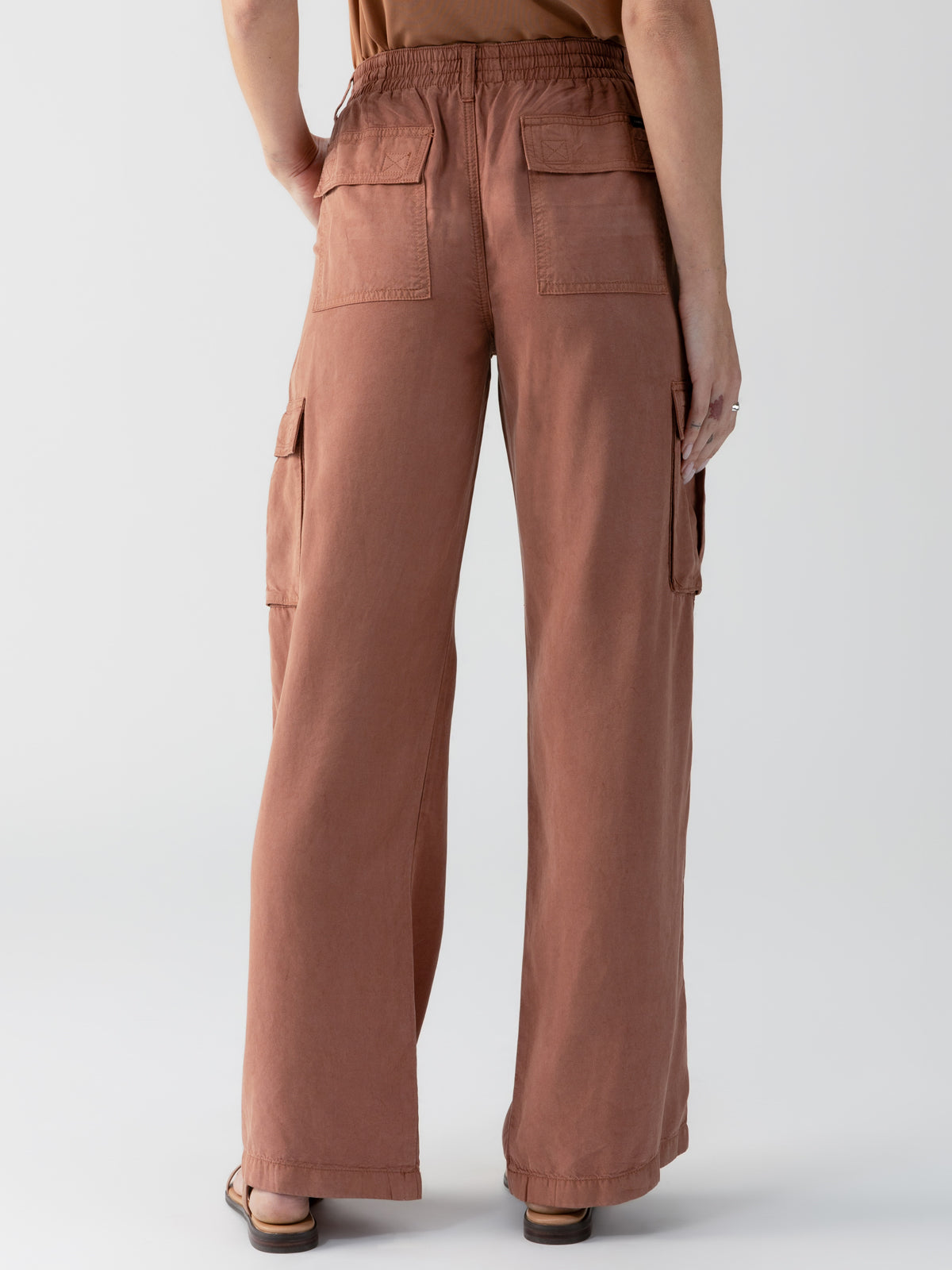 A person stands facing away, showcasing the Sanctuary Clothing Relaxed Reissue Washed Clay pants. These loose-fitting, high-waisted brown pants feature large rear pockets and side cargo pockets. They have an elastic waistband and are paired with sandals. The background is neutral and plain.