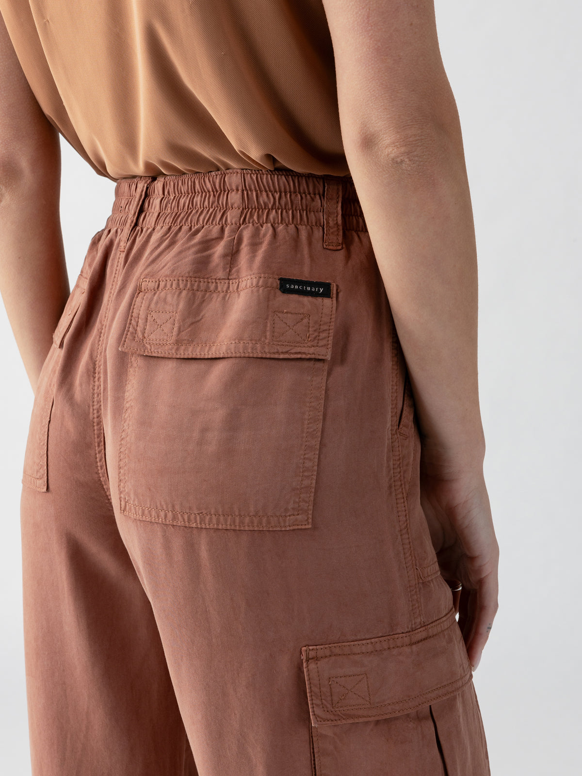 A person is shown from behind wearing the Relaxed Reissue Washed Clay pants by Sanctuary Clothing, featuring large back pockets and an elastic waistband. The right back pocket has a small black tag. The person is also wearing a matching light brown top against a plain, light-colored background.