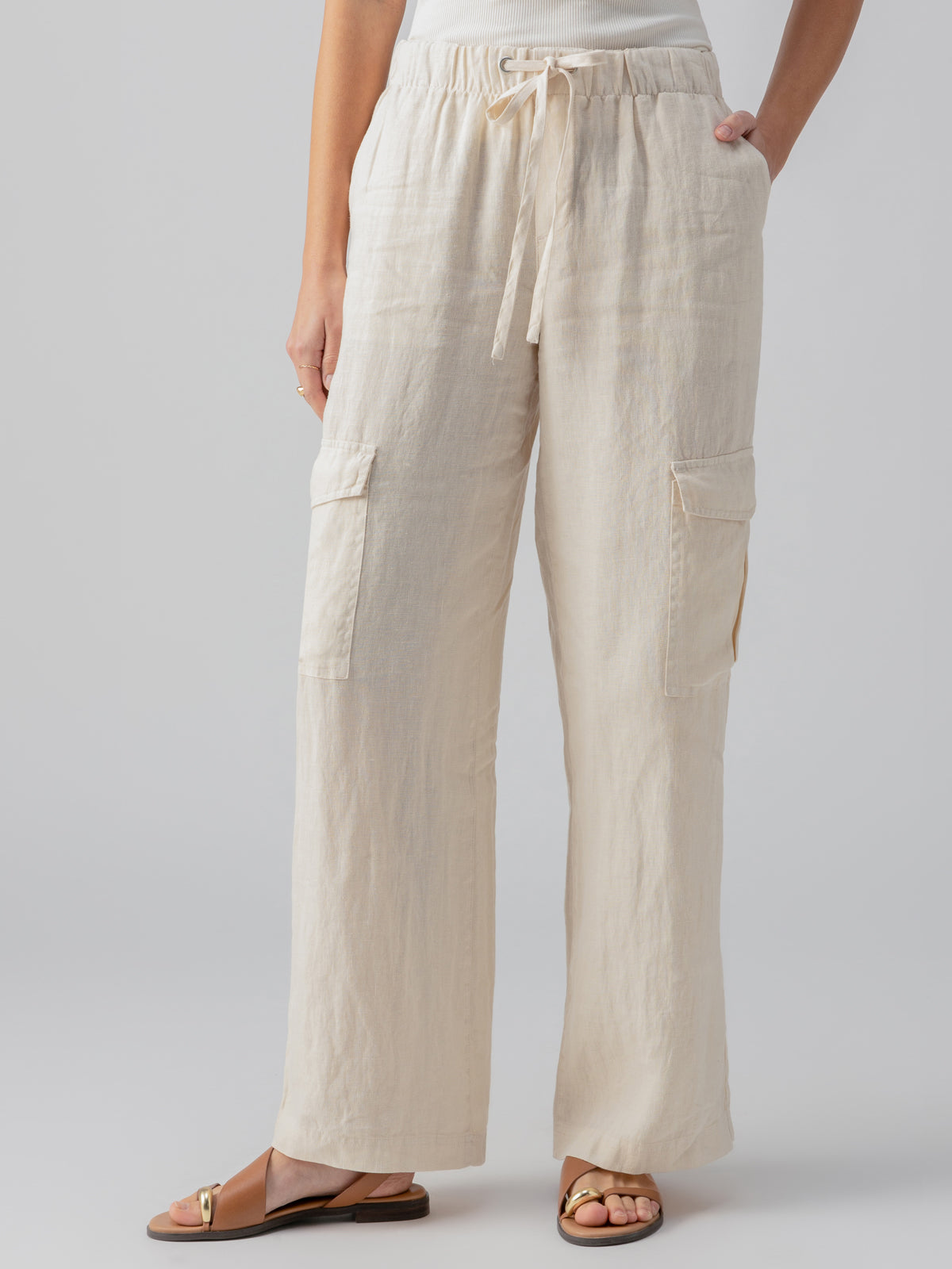 A person wearing the Drift Linen Cargo Birch pants by Sanctuary Clothing, which are beige, wide-legged with large pockets on the thighs and a drawstring waist, paired with a white top. One hand is placed in a pocket. The person is also wearing brown sandals with a gold buckle. The background is plain light grey.