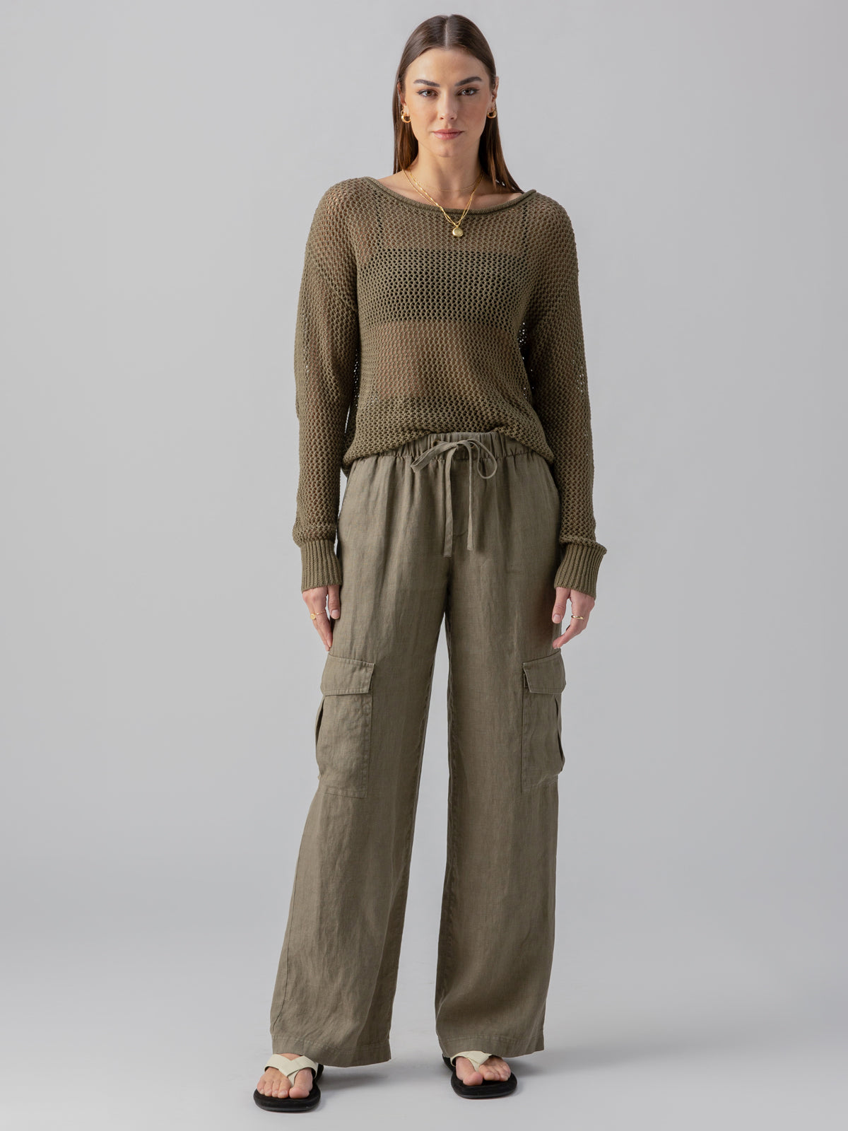 A person stands facing the camera, wearing an olive green, long-sleeved, knitted sweater over a light underlayer and loose-fitting "Drift Linen Cargo Burnt Olive" pants by Sanctuary Clothing, which feature side pockets. They have straight hair and are wearing sandals. The background is plain and light grey.