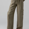 A person is wearing Drift Linen Cargo pants in Burnt Olive from Sanctuary Clothing that feature multiple pockets and a drawstring waistband. The person is also wearing a beige knit top and black sandals with white straps. The background is plain and light gray.