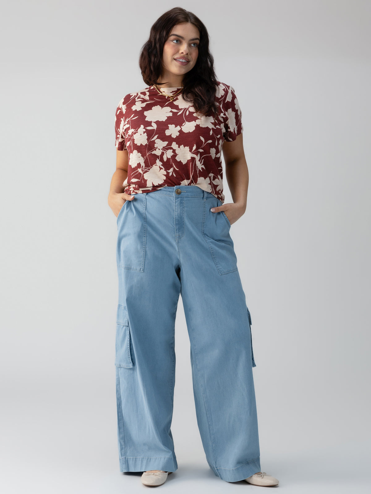 A woman with long dark hair is wearing a short-sleeved, red and white floral shirt paired with the Sunset Denim Pant Pale Blue from Sanctuary Clothing's Inclusive Collection and white shoes. She is standing with her hands in her pants pockets against a plain, light gray background.