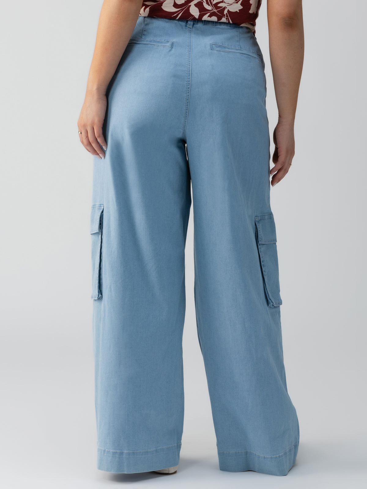 A person is standing with their back to the camera, wearing the Sunset Denim Pant in Pale Blue from Sanctuary Clothing's Inclusive Collection. The pants are wide-legged with pockets on the sides. They have a floral patterned top tucked into the pants. The background is plain and light-colored.