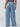 A person is standing with their back to the camera, wearing the Sunset Denim Pant in Pale Blue from Sanctuary Clothing's Inclusive Collection. The pants are wide-legged with pockets on the sides. They have a floral patterned top tucked into the pants. The background is plain and light-colored.