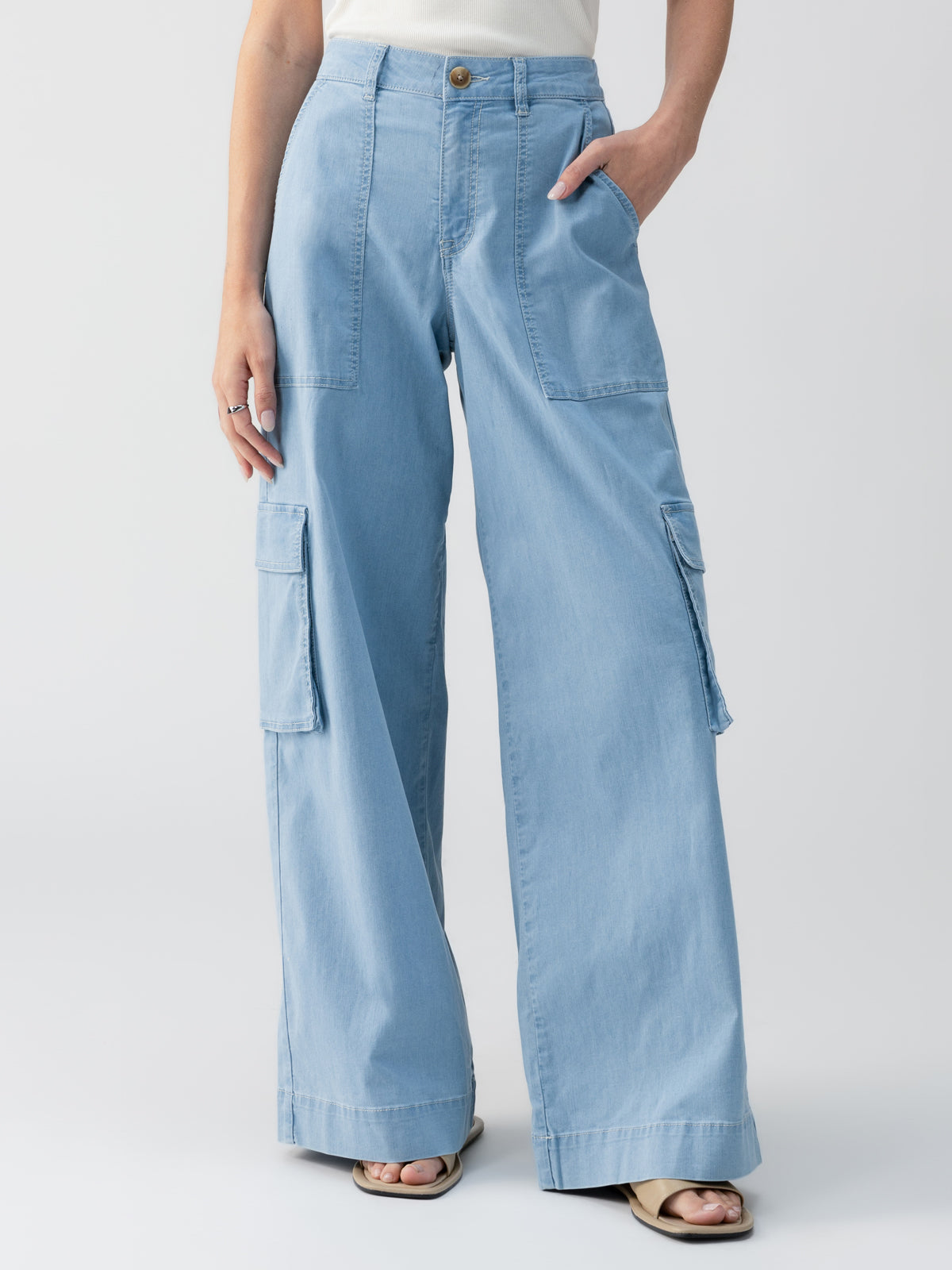 A person can be seen wearing the Sunset Denim Pant Pale Blue by Sanctuary Clothing. This light blue, high-waisted, wide-leg cargo pants feature large pockets on the thighs. The individual is posing with one hand in their pocket, paired with a simple, tucked-in top and sandals against a plain white background.