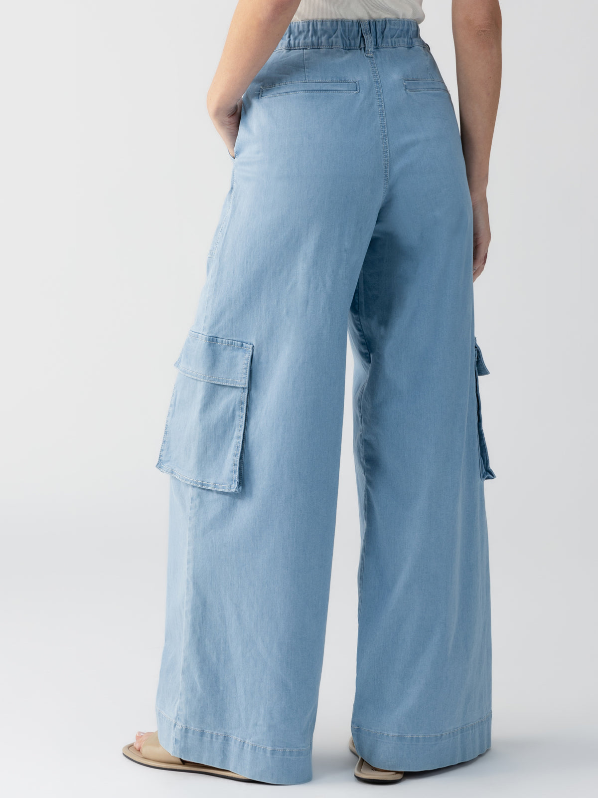 A person is shown from the back, wearing the Sanctuary Clothing Sunset Denim Pant in Pale Blue, which features wide-legs and large side pockets. They are also wearing a light-colored top tucked into the jeans. The background is plain white.