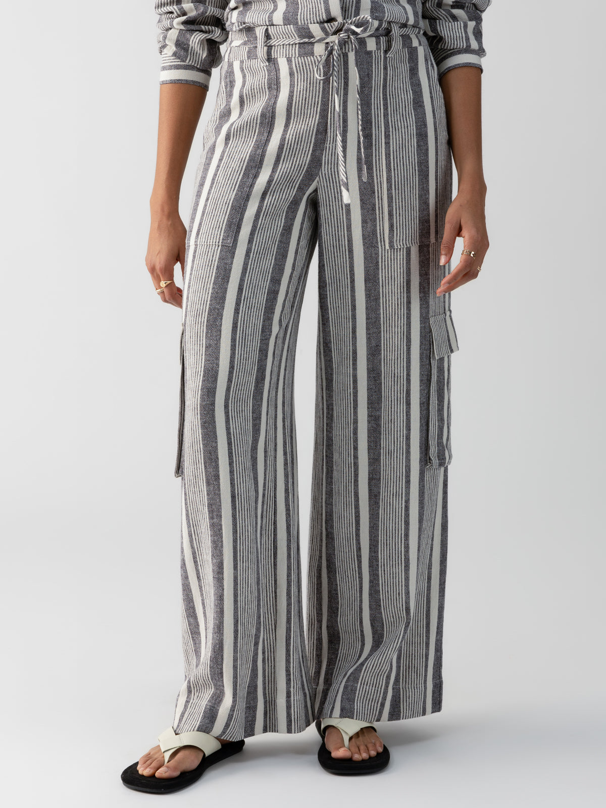 A person is wearing the Sunset Linen Pant Varigated Stripe from Sanctuary Clothing, featuring high-waisted, wide-leg pants with vertical gray and white stripes. The pants are cinched at the waist with a matching fabric belt. The person pairs these stylish pants with a striped long-sleeve shirt and black and white slip-on sandals against a plain white background.