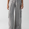 A person is wearing the Sunset Linen Pant Varigated Stripe from Sanctuary Clothing, featuring high-waisted, wide-leg pants with vertical gray and white stripes. The pants are cinched at the waist with a matching fabric belt. The person pairs these stylish pants with a striped long-sleeve shirt and black and white slip-on sandals against a plain white background.