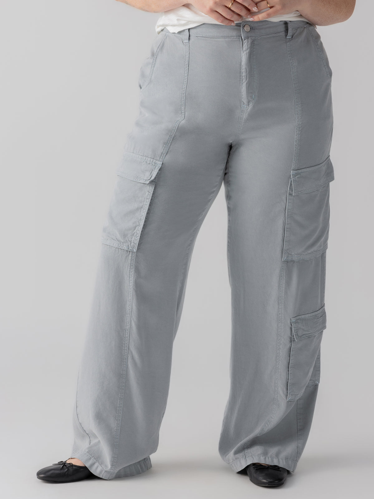 A person is wearing the Doheny Cargo Eucalyptus pants from the Inclusive Collection by Sanctuary Clothing and a white top. They are standing in a neutral pose, showing the front of the pants, and are wearing black flat shoes. The background is plain and gray.