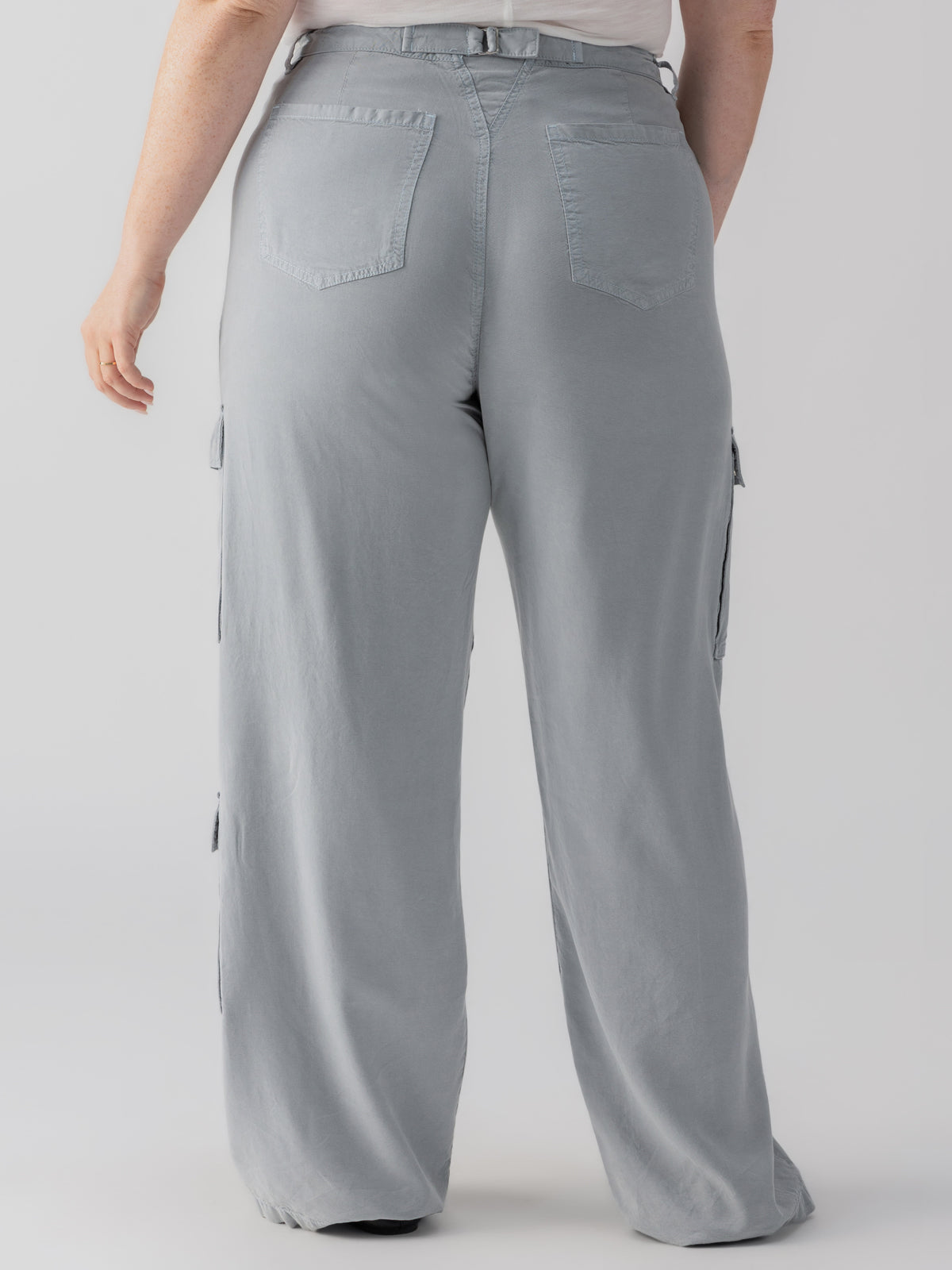 A person wearing the Doheny Cargo Eucalyptus Inclusive Collection pants by Sanctuary Clothing, which are light grey with wide legs and large pockets on the sides and back, is facing away from the camera. The person is also wearing a white shirt. The background is plain and neutral.