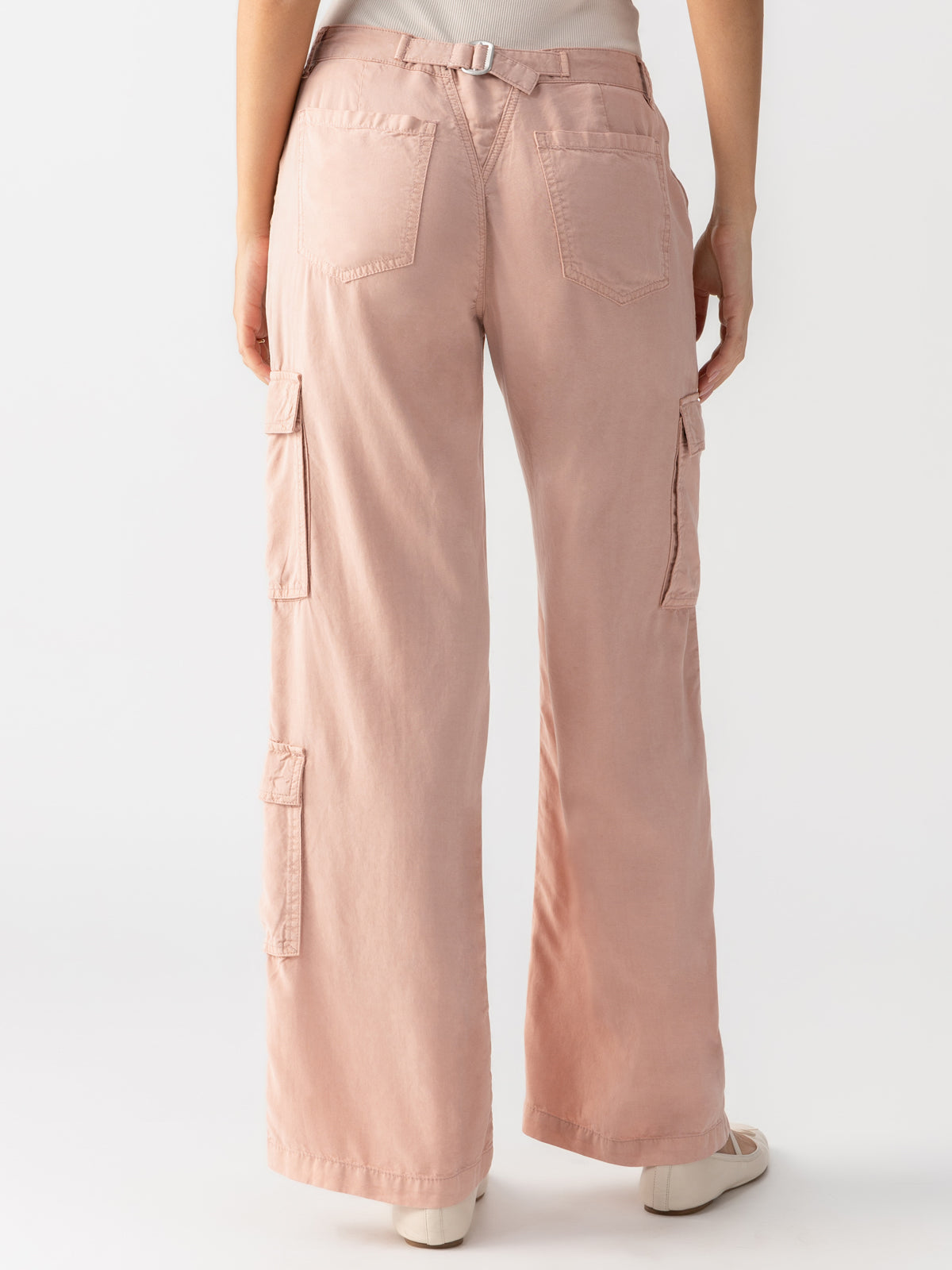 A person is standing with their back to the camera, wearing Sanctuary Clothing's Doheny Cargo Smokey Rose pants in a light pink color, featuring multiple side and back pockets. The loose-fitting, cuffed-at-the-bottom cargo pants are paired with white sneakers against a plain white background.