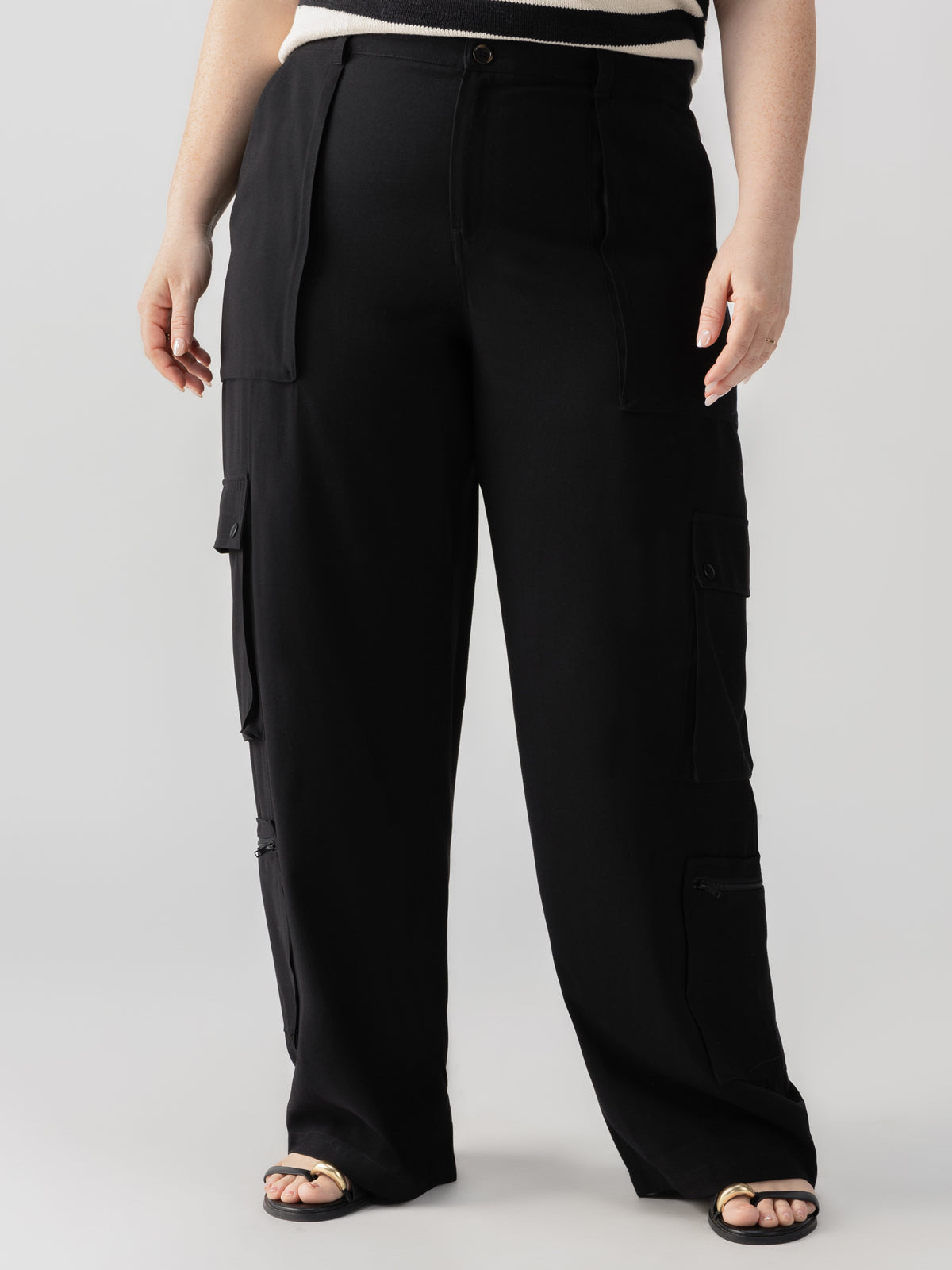 A person wearing the Ott Cargo Black Inclusive Collection trousers from Sanctuary Clothing and a black and white top stands against a neutral background. The trousers feature multiple pockets on the legs, and the individual is also wearing open-toe sandals.