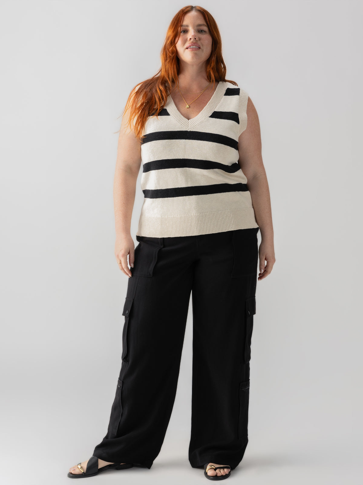 A woman with long red hair is wearing a sleeveless, V-neck white sweater with black horizontal stripes and the Ott Cargo Black Inclusive Collection pants from Sanctuary Clothing. She has on black sandals and is standing against a plain white background.