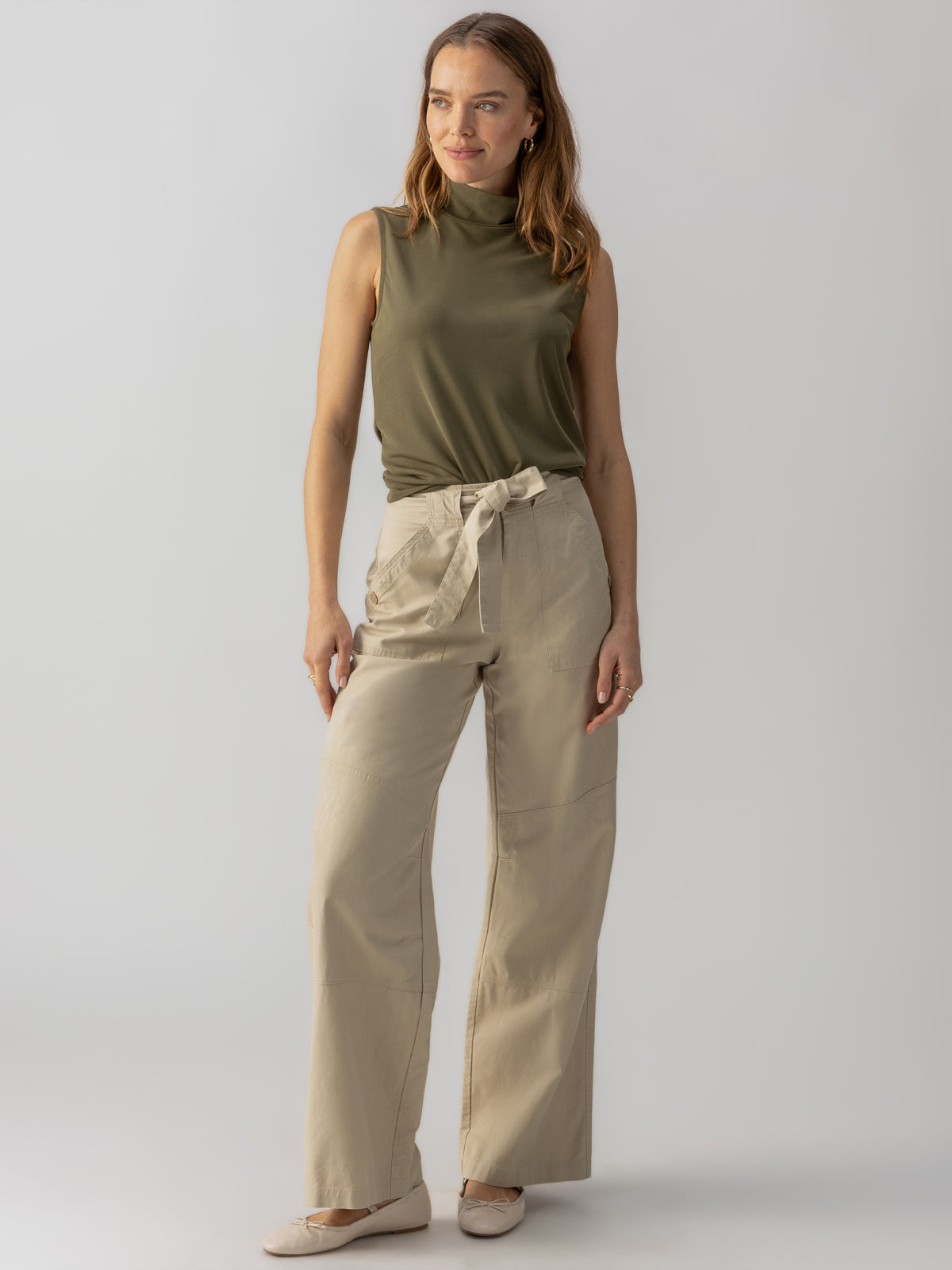 A woman stands confidently against a plain background wearing a sleeveless olive green turtleneck, Sanctuary Clothing's Reissue 90S Sash Extended Marble Beige high-waisted wide-leg trousers with a tie belt, and beige flats. She has long wavy hair and a gentle smile on her face.