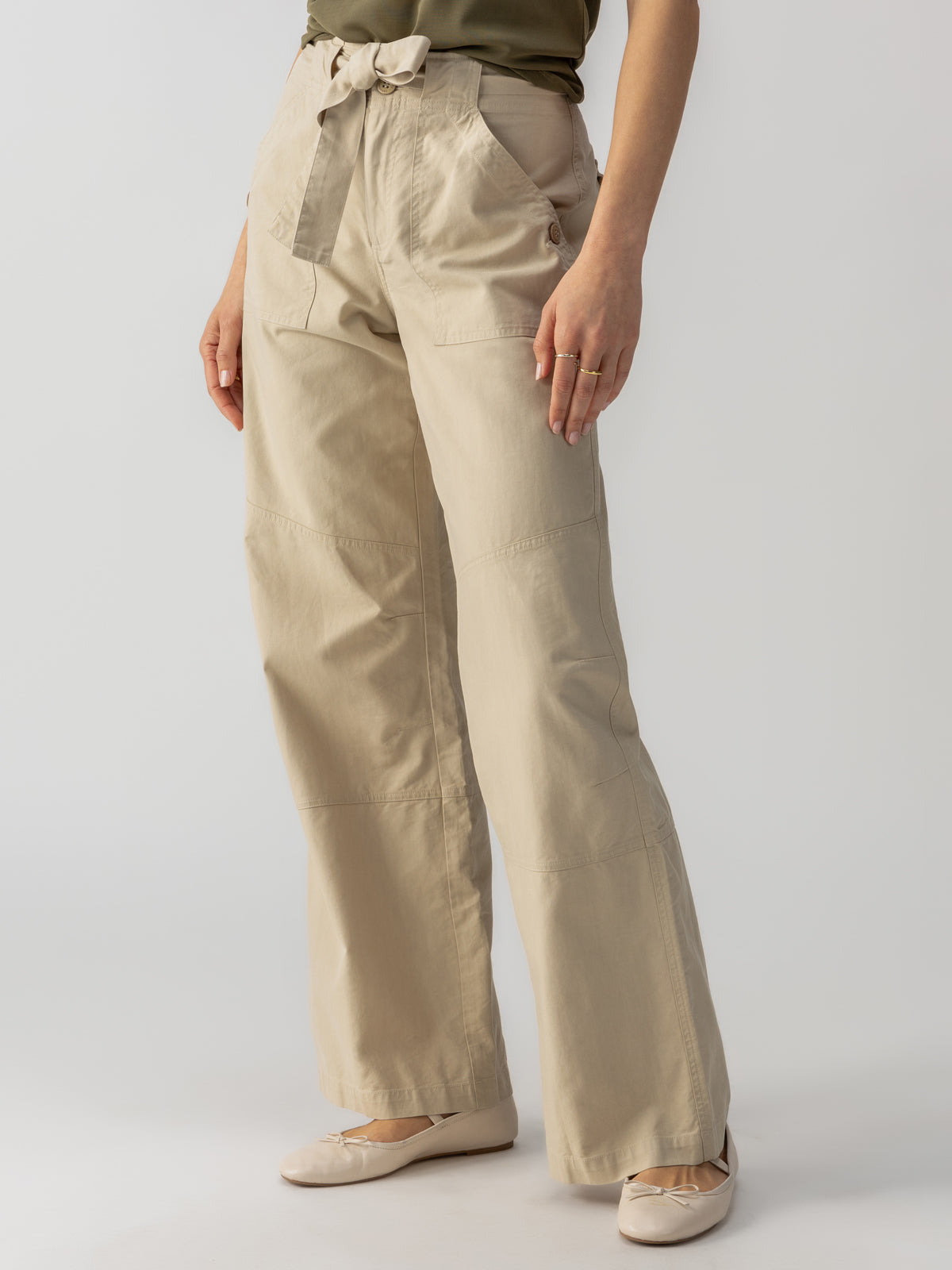 The individual is adorned in Sanctuary Clothing's Reissue 90s Sash Extended Marble Beige high-waisted, wide-leg trousers with a tie belt, paired with a green top. They are standing confidently with hands in pockets, showcasing the casual and comfortable ensemble, completed by light-colored flat shoes.