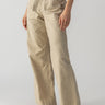 The individual is adorned in Sanctuary Clothing's Reissue 90s Sash Extended Marble Beige high-waisted, wide-leg trousers with a tie belt, paired with a green top. They are standing confidently with hands in pockets, showcasing the casual and comfortable ensemble, completed by light-colored flat shoes.