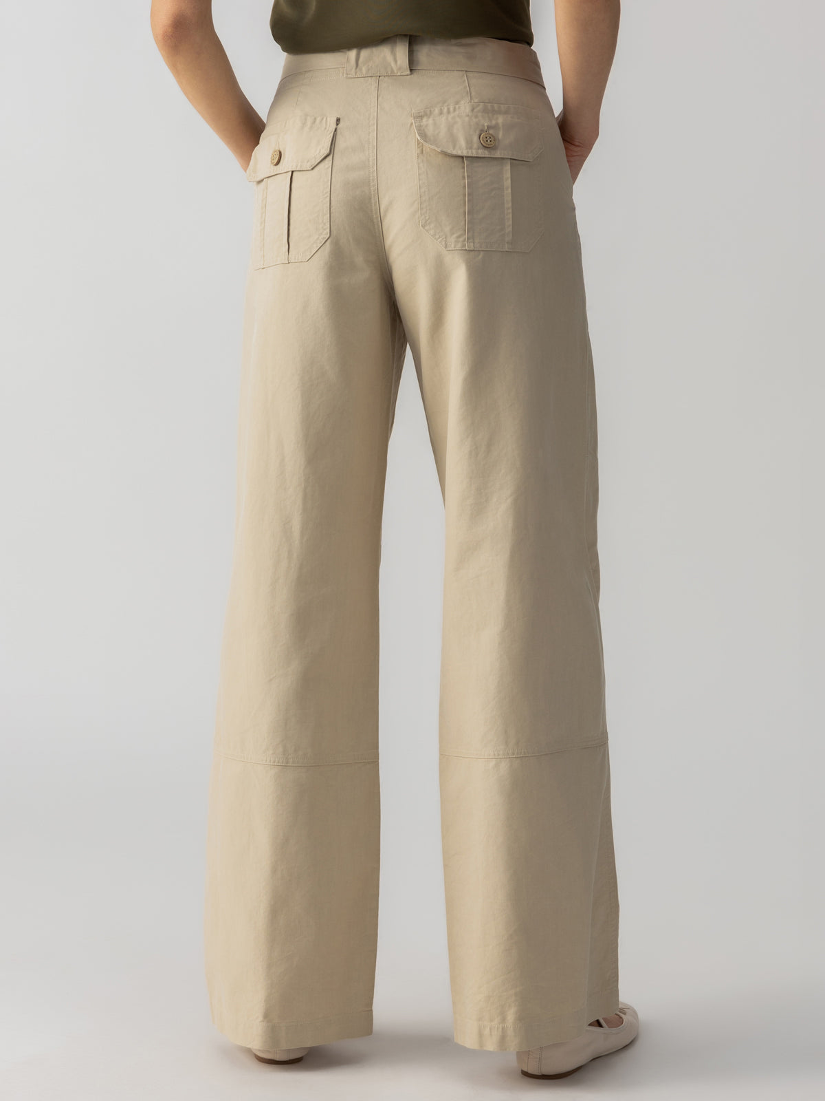 Rear view of a person wearing the Sanctuary Clothing Reissue 90S Sash Extended Marble Beige wide-leg pants with large patch pockets on the back. The high-waisted pants are paired with a tucked-in olive green top. The person stands, showcasing the full length of the pants against a plain light-colored background.