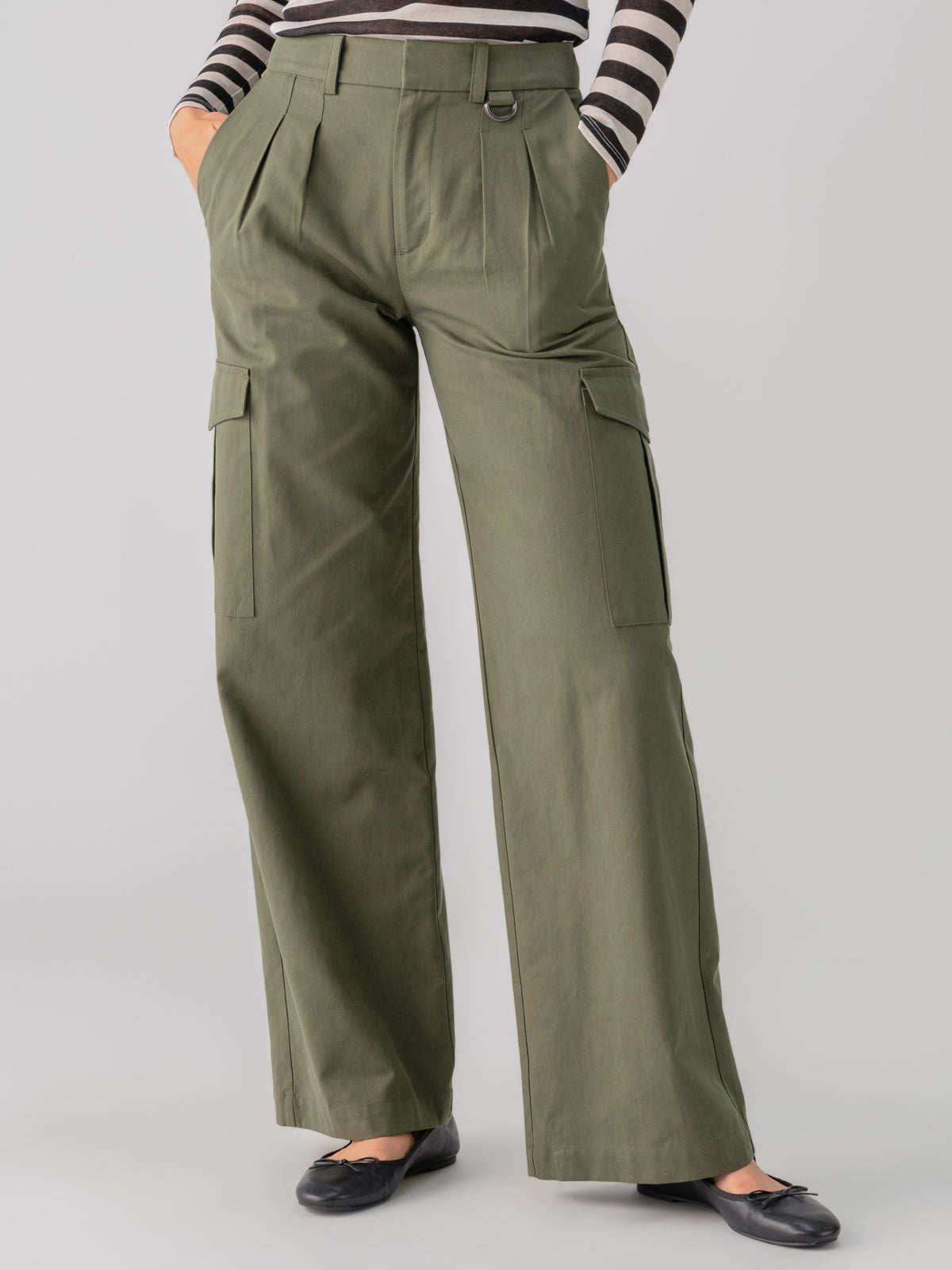 A person is wearing the Frankie Cargo Dark Olive pants from Sanctuary Clothing, which feature wide legs, pleats, side pockets, and flap pockets on the thighs. They are also wearing a black and white striped long-sleeve shirt and black ballet flats. The background is plain gray.