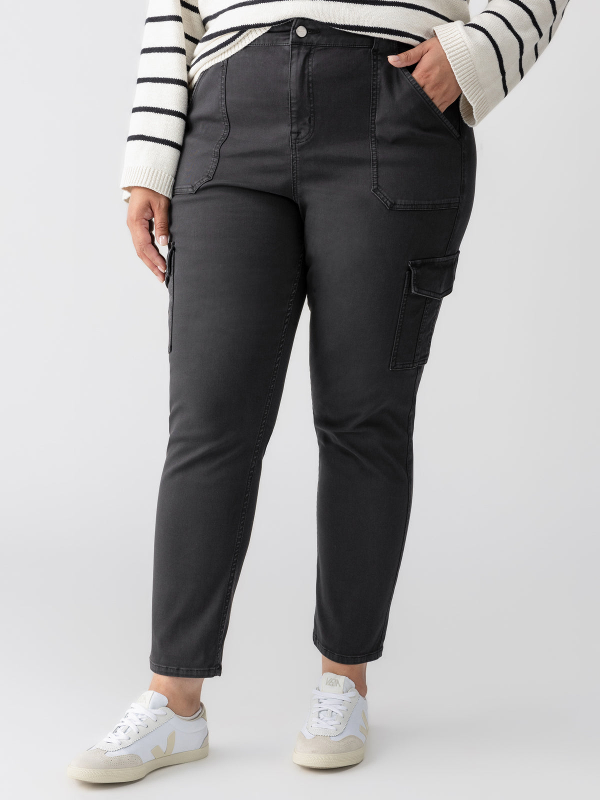 A person is wearing the Sculpted Hayden Cargo Black pants from Sanctuary Clothing's Inclusive Collection, paired with a white and black striped sweater. Their right hand is placed in their pants pocket, and they are wearing white sneakers. The background is plain white.