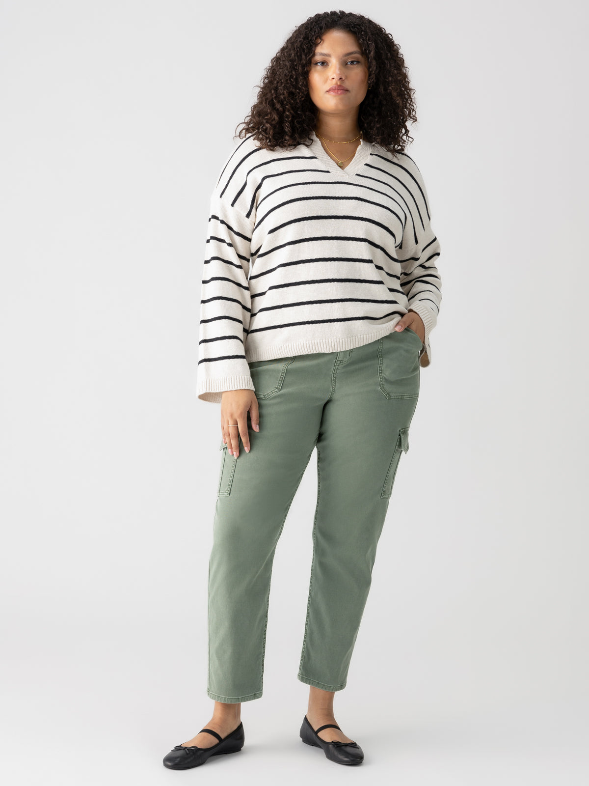 A person with curly hair stands against a plain background, wearing a cream-colored sweater with black stripes, Sanctuary Clothing's Sculpted Hayden Cargo Dark Spruce Inclusive Collection pants, and black flats. Their hands are loosely placed in their pants pockets.