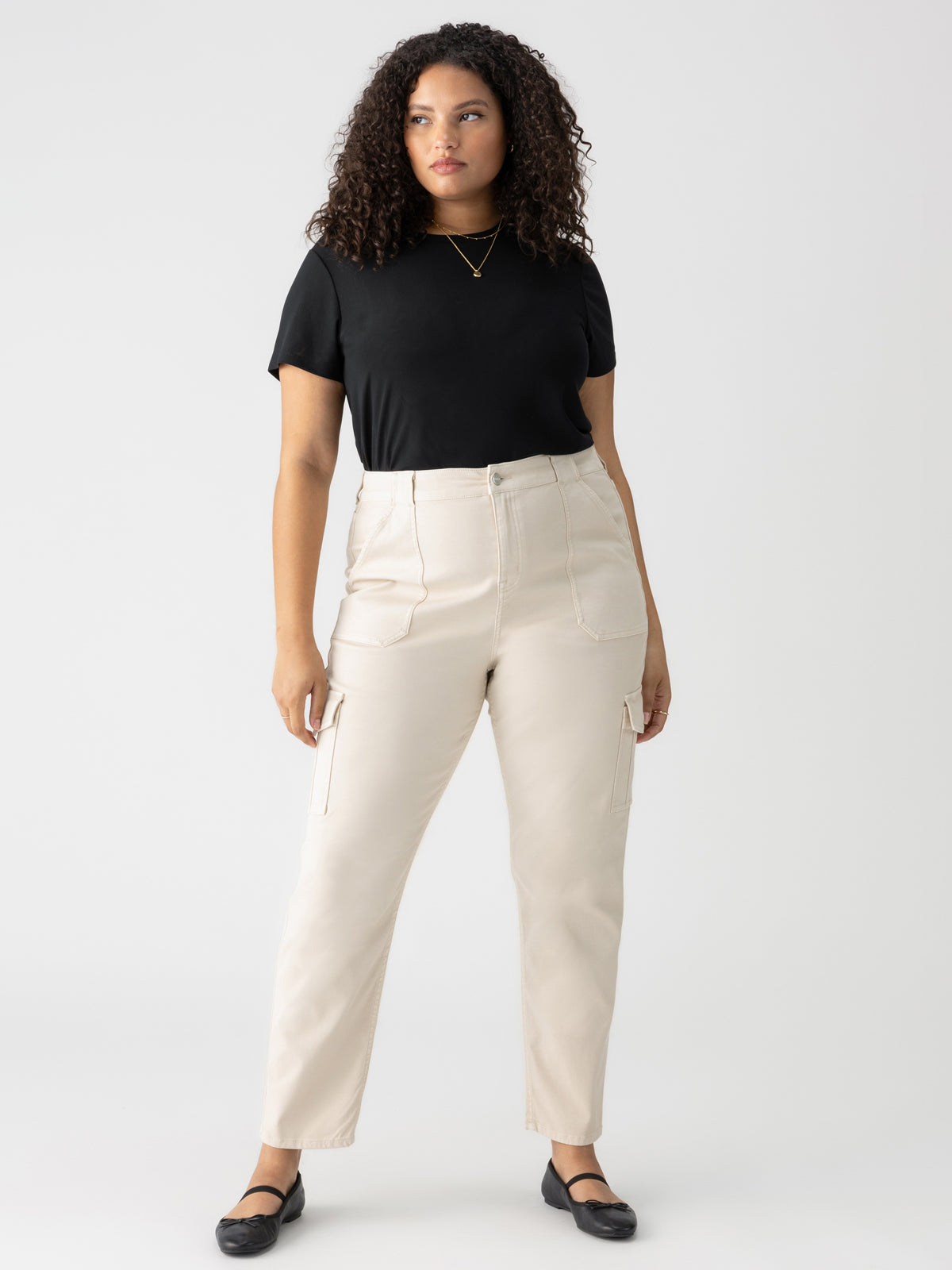 A person with curly hair is standing against a plain white background. They are wearing a black t-shirt, the Sculpted Hayden Cargo pants in Toasted Almond from the Inclusive Collection by Sanctuary Clothing, and black flats. They appear to be looking slightly to the side with a calm expression.