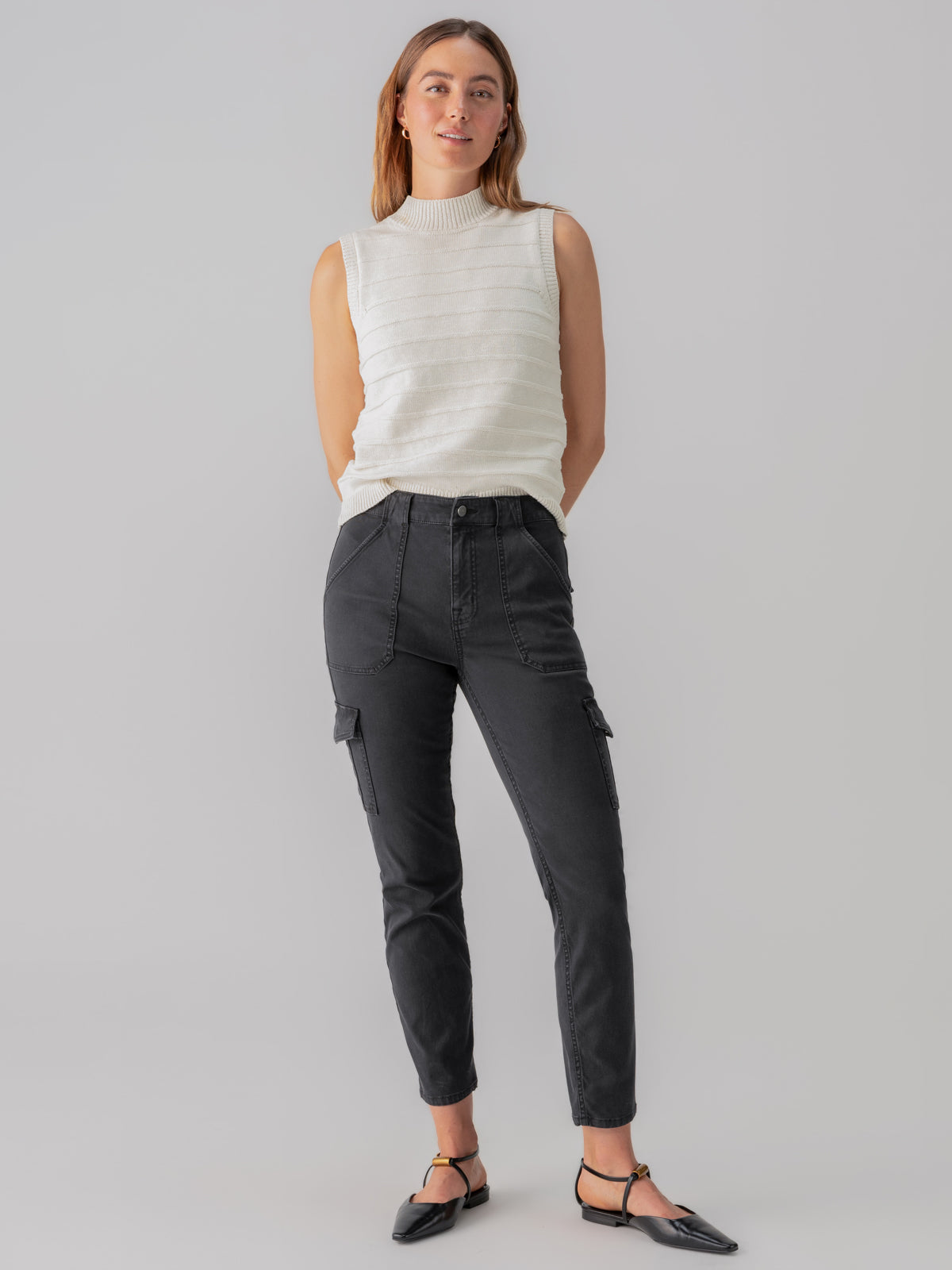 A person stands against a plain backdrop, wearing a sleeveless white knit top and Sanctuary Clothing's Sculpted Hayden Cargo Black pants. They have shoulder-length hair, are looking slightly to the side, with their hands resting behind their back, and are wearing black pointed shoes.