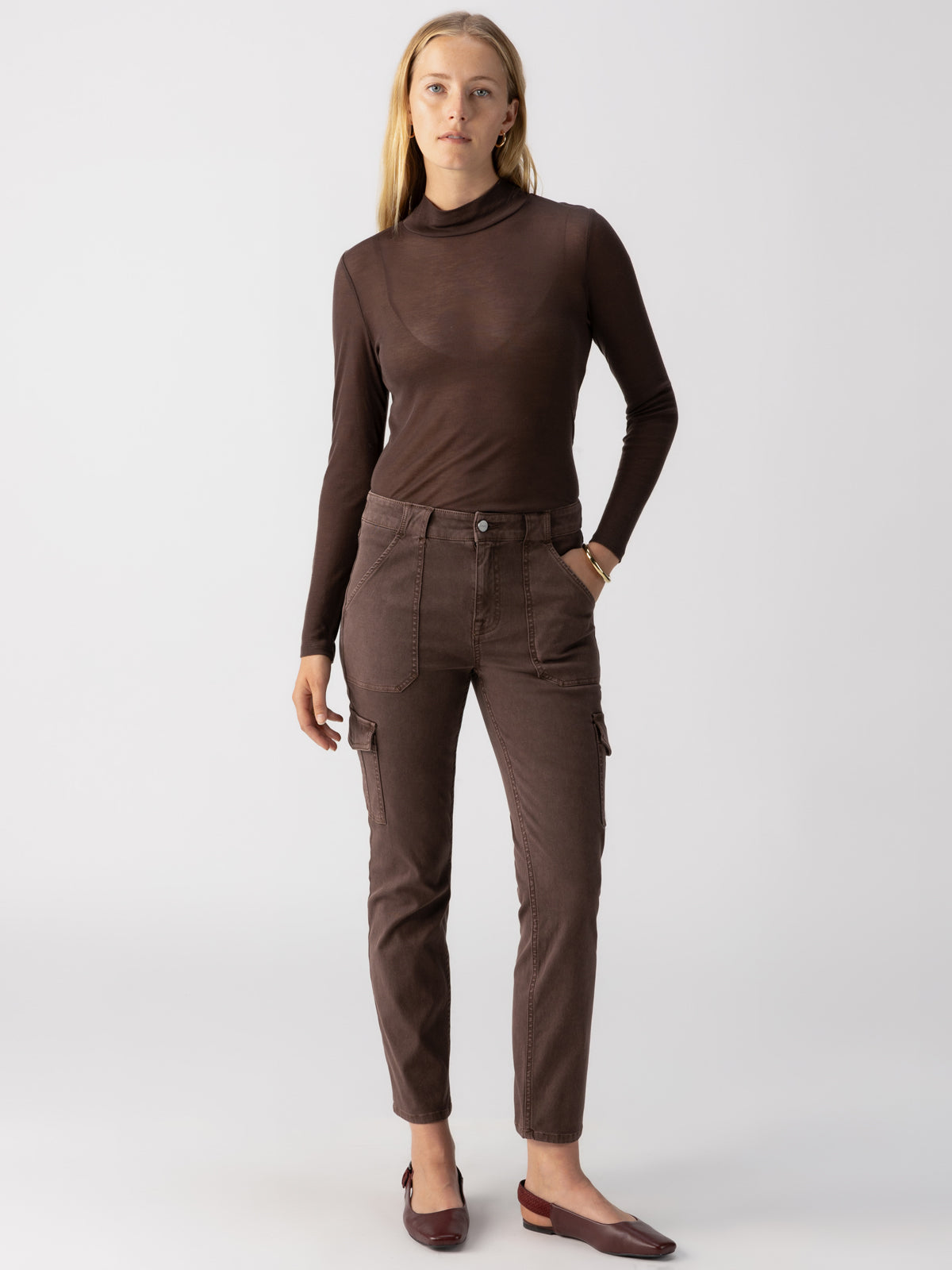 A person with long blonde hair is standing against a plain background, wearing a brown long-sleeve turtleneck, matching Sanctuary Clothing Sculpted Hayden Cargo Brown Sugar pants, and brown loafers. They have a neutral expression and their arms are relaxed by their sides.