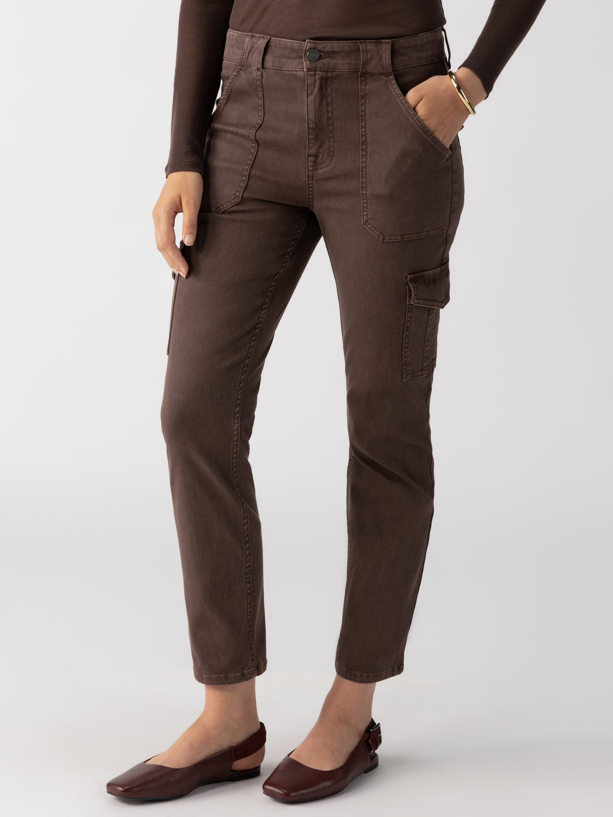 A person wearing the Sculpted Hayden Cargo Brown Sugar pants by Sanctuary Clothing is shown from the shoulders down against a plain background. The high-waisted brown cargo pants feature front and side pockets. They are also wearing a long-sleeved brown top and brown flats. One hand is placed in a pocket, while the other rests by their side.