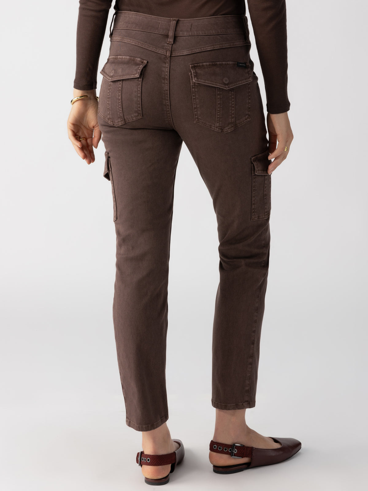 A person standing and facing away from the camera, wearing the Sanctuary Clothing Sculpted Hayden Cargo Brown Sugar pants with multiple pockets, including back and side cargo pockets. They are also wearing brown shoes and a matching brown long-sleeve top.