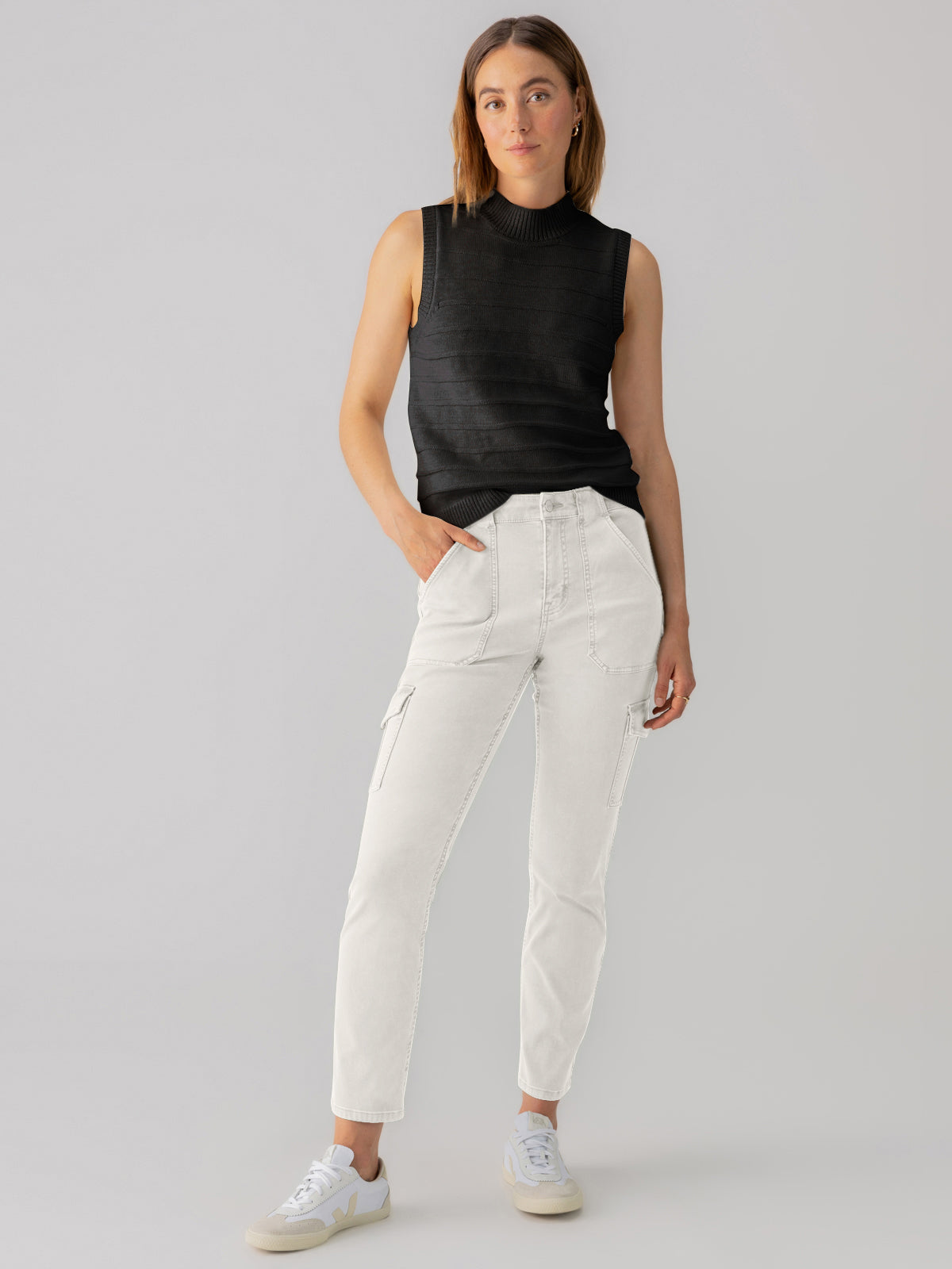 A person stands confidently in a sleek, sleeveless black top paired with the Sanctuary Clothing's sculpted hayden cargo pants in pumice stone, their hands comfortably resting in the pockets. Casual sneakers complete the look as they maintain a neutral expression against a plain gray background.