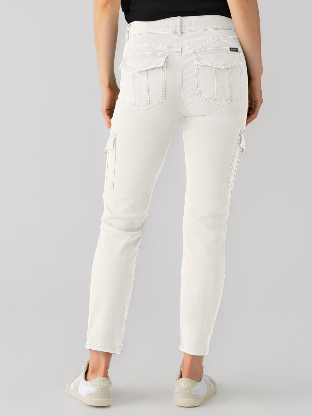 A person is shown from the rear wearing the "sculpted hayden cargo pumice stone" jeans by Sanctuary Clothing, paired with a black top. The jeans feature multiple pockets and are complemented by white sneakers, set against a plain light gray background.