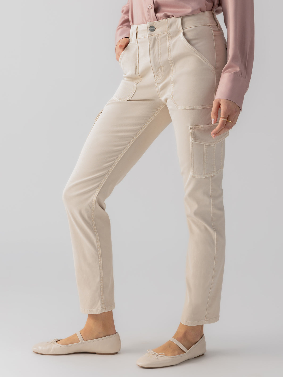 A person stands angled sideways, showcasing the Sculpted Hayden Cargo Toasted Almond pants by Sanctuary Clothing. The beige high-waisted straight-leg pants feature large front pockets and side cargo pockets. They wear a dusty pink long-sleeve blouse tucked into the pants and beige flat shoes with bows. The background is a solid light color.