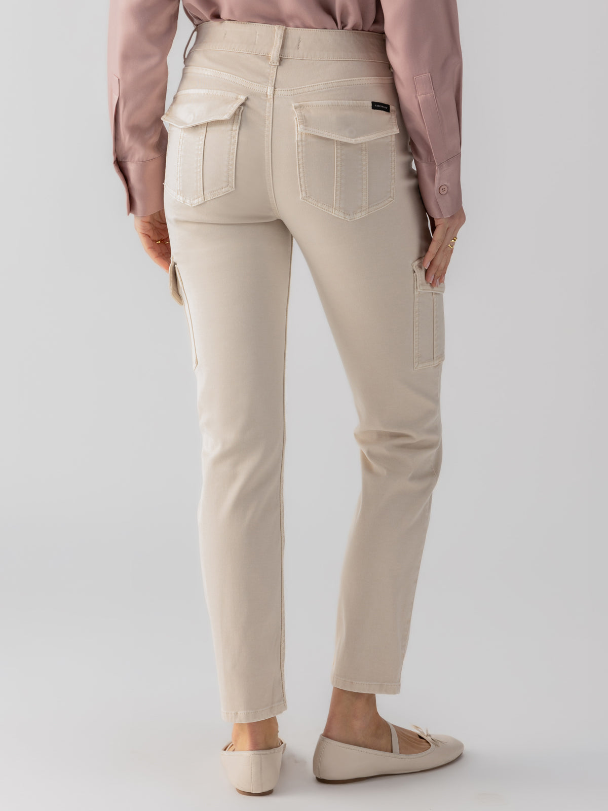 A person is seen from behind, wearing a long-sleeved mauve shirt and a pair of beige Sanctuary Clothing's Sculpted Hayden Cargo Toasted Almond pants, which feature large back and side pockets. The person is also wearing beige, flat-soled shoes. The background is plain and light-colored.