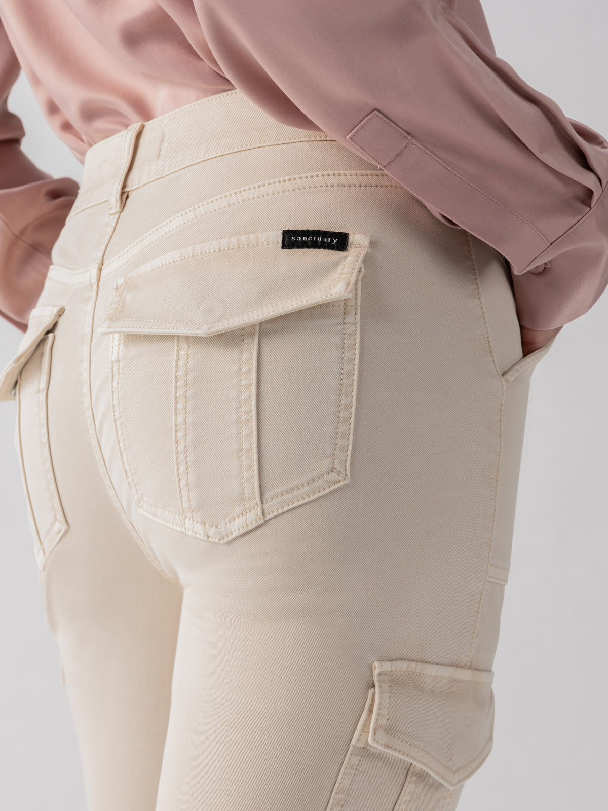 A person wearing the Sanctuary Clothing Sculpted Hayden Cargo pants in Toasted Almond and a pink long-sleeved shirt. The focus is on the back pocket area of the pants, highlighting the pocket design and stitching details. A black label with text is sewn onto one of the pockets.