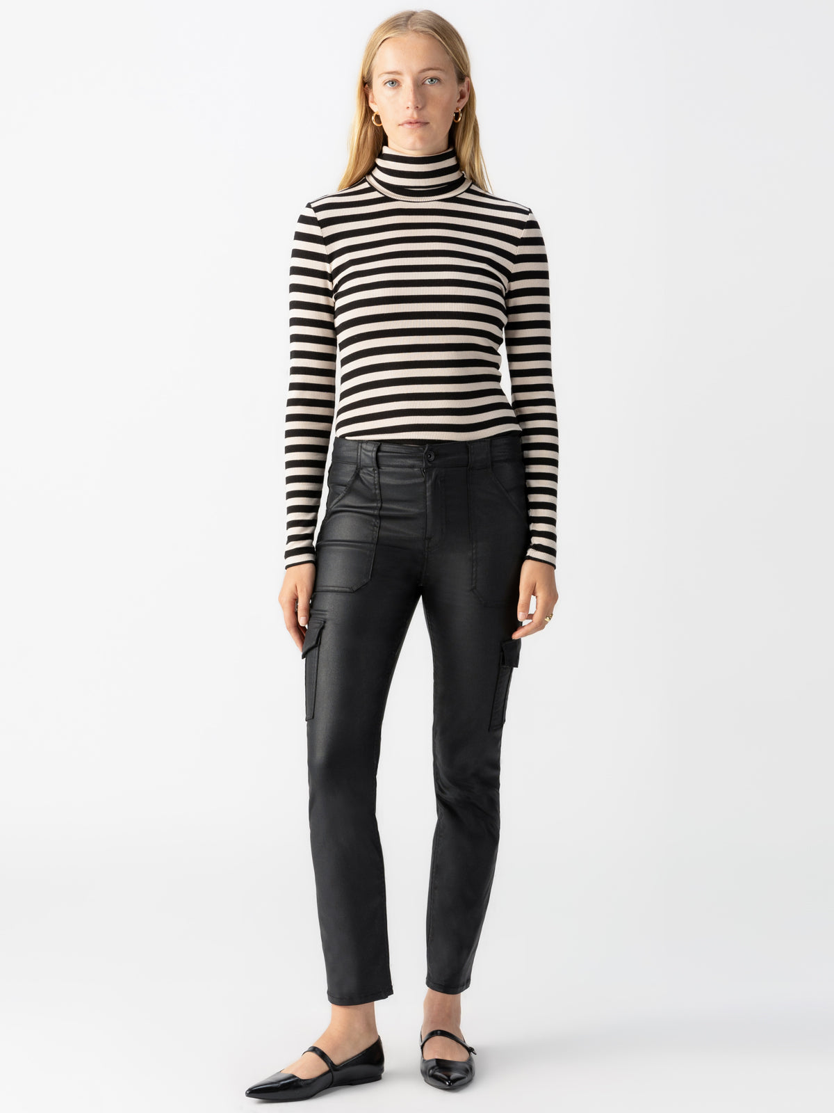 A woman stands facing forward against a plain white background. She is wearing a black and white striped long-sleeve turtleneck, Sanctuary Clothing's Sculpted Hayden Cargo Slick Black fitted pants with front pockets, and black pointed-toe shoes. She has straight hair and a neutral expression.