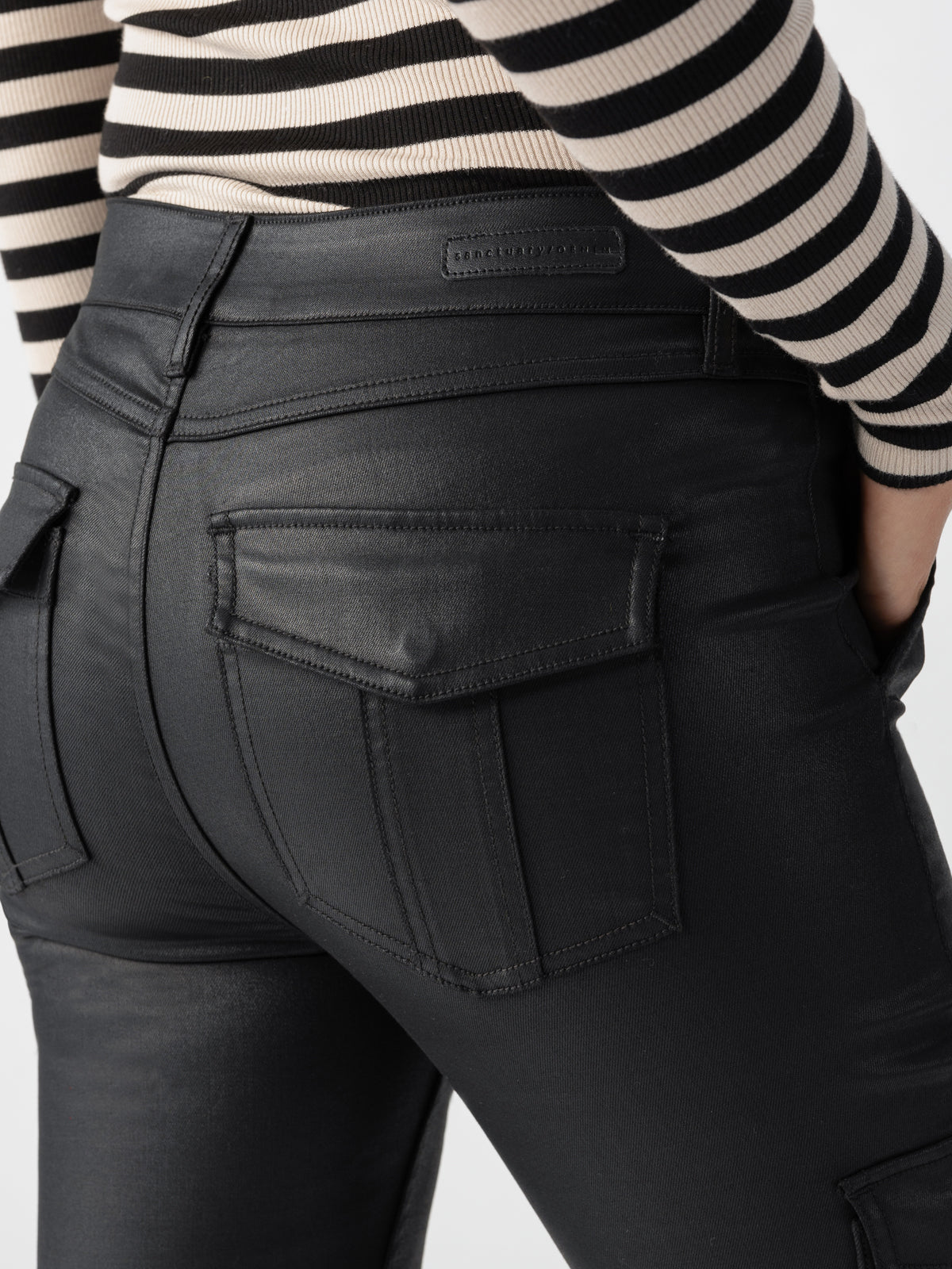 A person wearing Sculpted Hayden Cargo Slick Black pants by Sanctuary Clothing, featuring visible pocket details, paired with a black and white striped shirt. The focus is on the back pockets of the pants, with the person's hand partially visible, tucked into one of the side pockets.