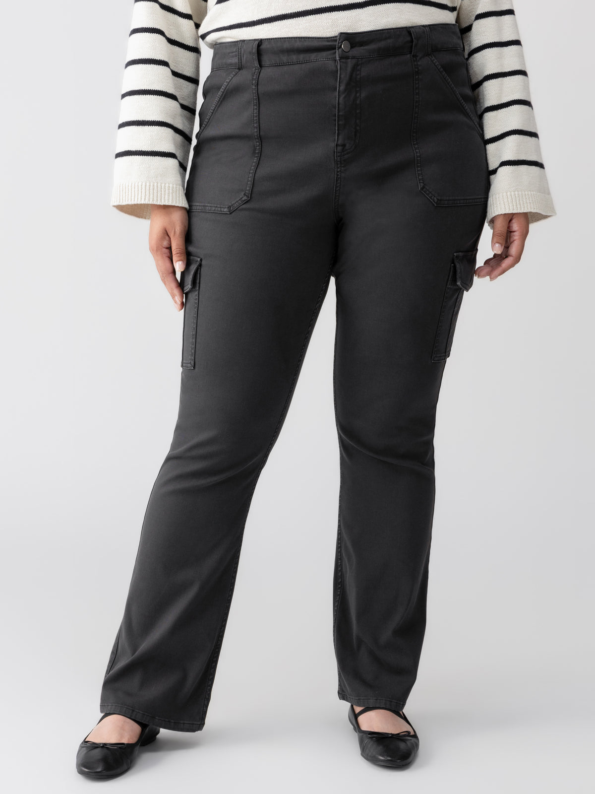 A person stands wearing Sanctuary Clothing's Sculpted Hayden Bootcut Black Inclusive Collection pants, which feature front and side pockets, paired with a white sweater adorned with black horizontal stripes. The person’s feet are in black flat shoes, and the background is a plain light gray.