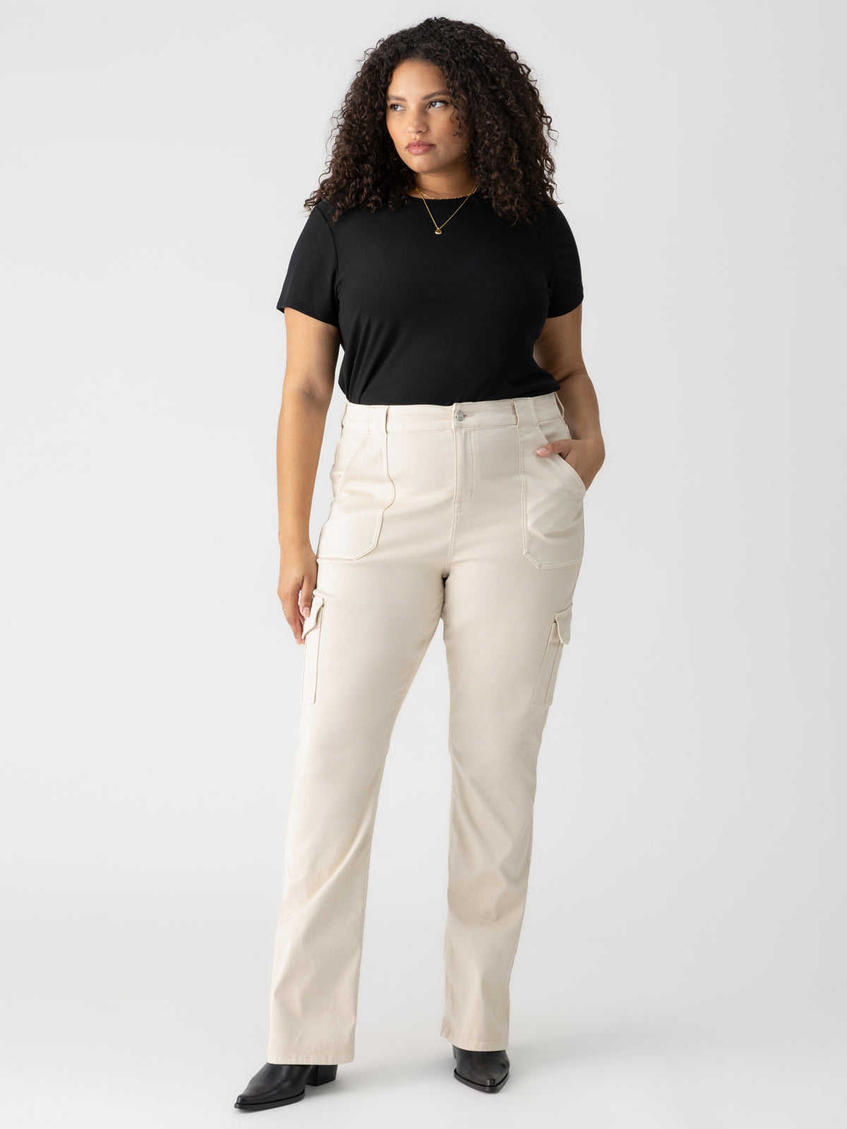 A woman with curly hair is wearing a black short-sleeve top, light beige high-waisted cargo pants with pockets, and the Sculpted Hayden Bootcut Toasted Almond boots from Sanctuary Clothing. She stands facing slightly to the right against a plain white background. She is also wearing a gold necklace with a small pendant.