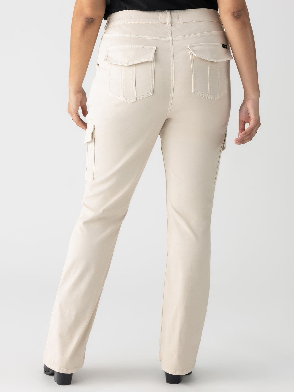 A person wearing the Sculpted Hayden Bootcut Toasted Almond pants from Sanctuary Clothing's Inclusive Collection stands with their back to the camera. The beige cargo pants feature multiple pockets, including two large flap pockets on the back and side pockets on the thighs. The person wears black shoes, and the background is plain and light-colored.