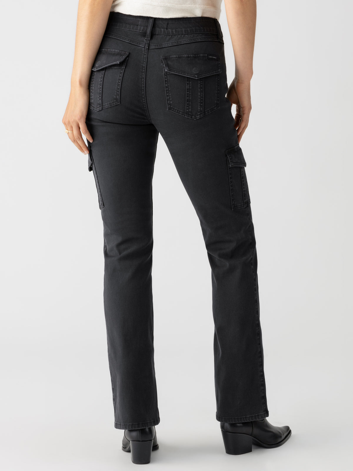 A person is shown from the back, wearing a pair of black cargo jeans with flap pockets and seams, and a light-colored top. The person is also wearing a pair of Sculpted Hayden Bootcut Black heeled boots by Sanctuary Clothing. The background is plain and light-colored.